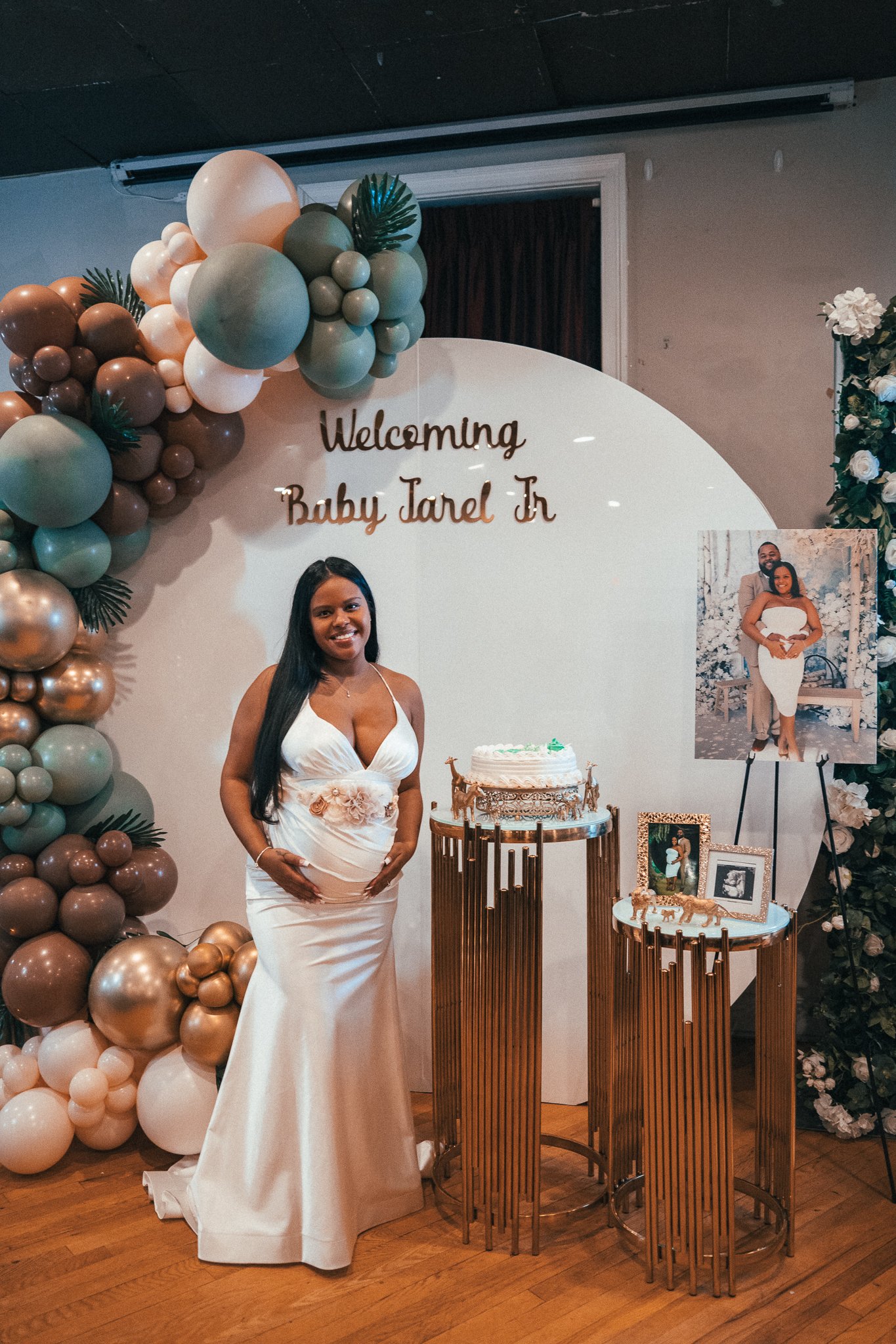 A woman in an elegant white gown standing in front of a birthday celebration backdrop with balloons and framed photos, with a sign reading 'Welcoming Baby Jarel Jr'.