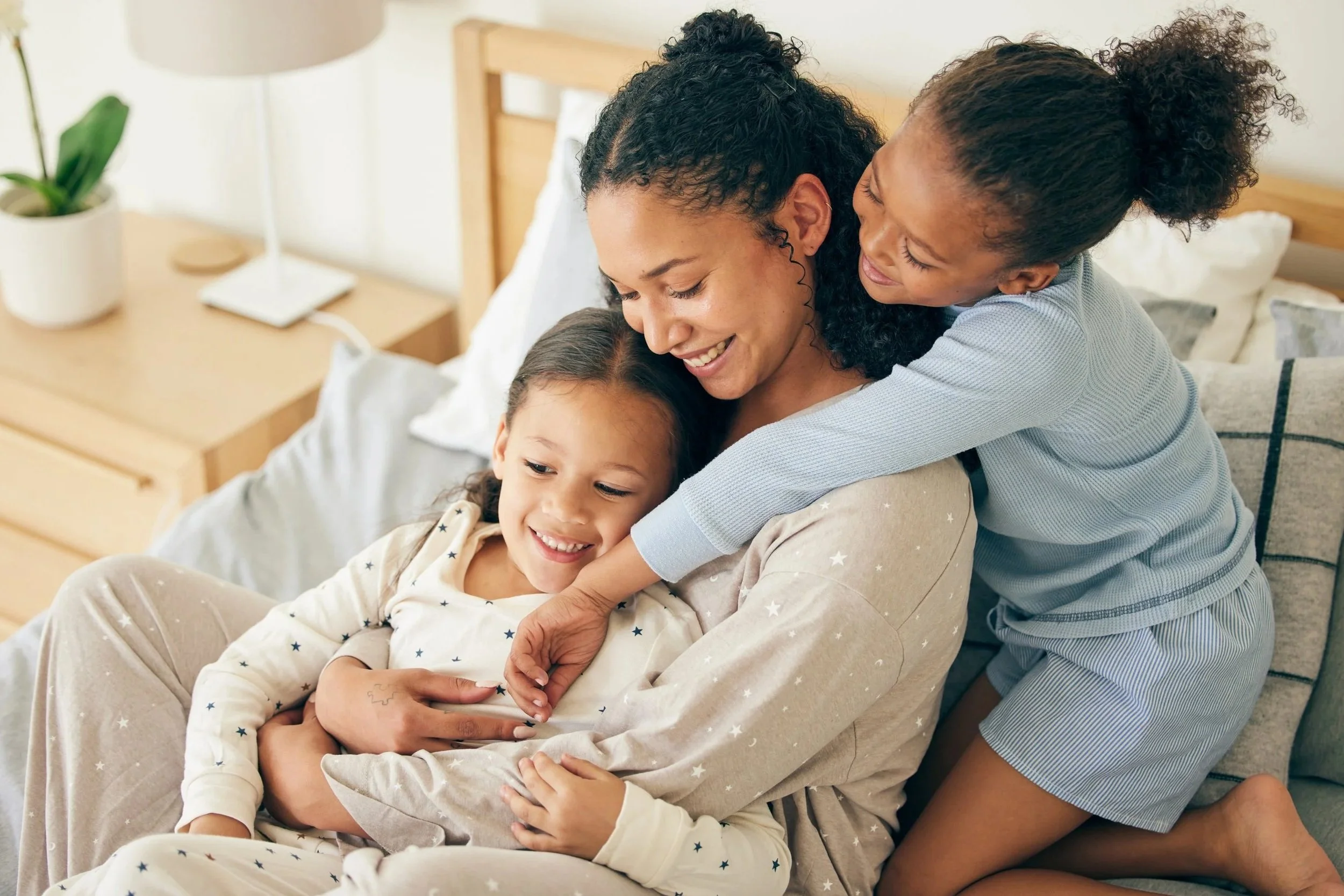 A woman with curly black hair sits on a bed, smiling and hugging her two daughters who are also smiling and embracing her. The daughters have curly hair, and the room is softly lit with a bedside table, a lamp, and a plant visible in the background.