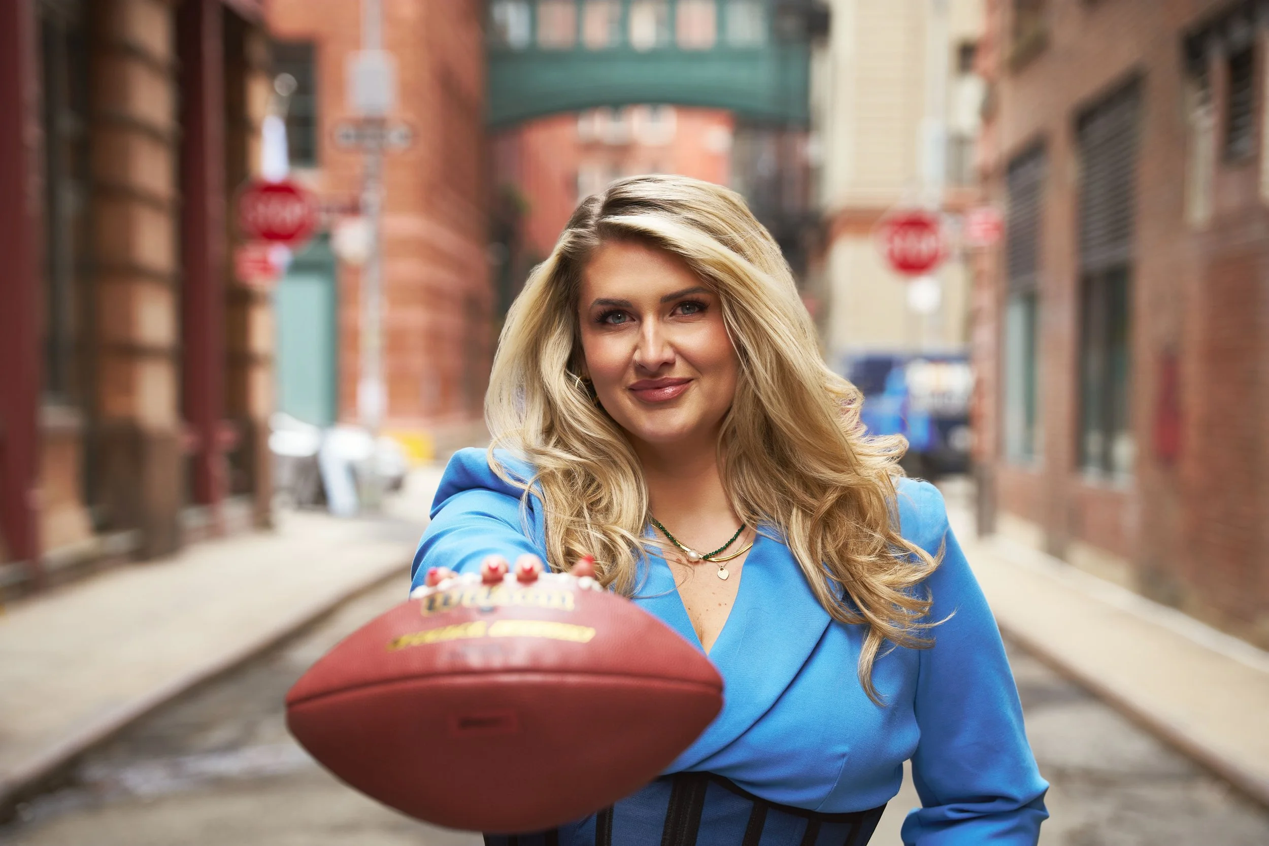 A woman in a blue blazer reaching out towards the camera with a football, on a city street with brick buildings and stop signs in the background.