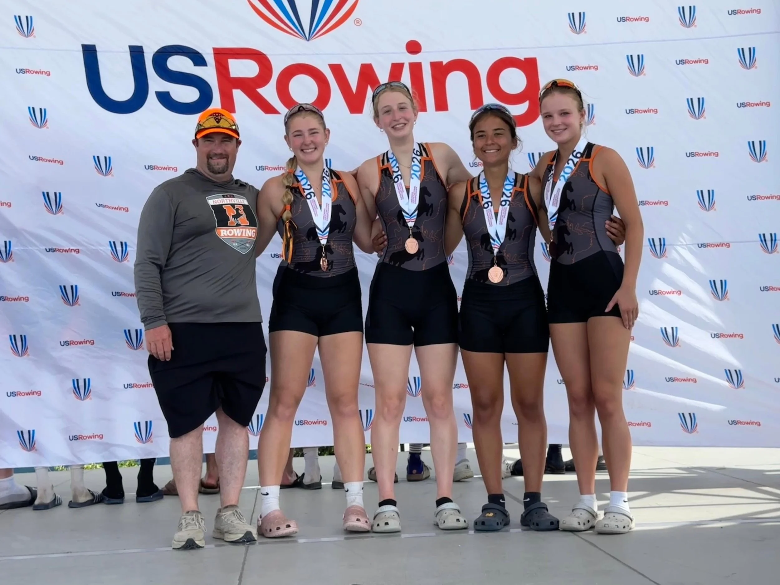 Group of five people, four young women and one man, standing together in front of a U.S. Rowing banner after a rowing event. The women are wearing athletic outfits with medals around their necks, indicating they are winners. The man, standing on the far left, is dressed in casual athletic clothing and a cap. They are smiling and appear to be celebrating their achievement.