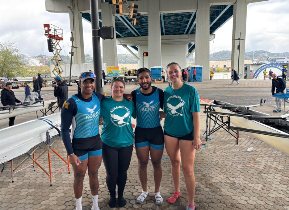 Four rowers standing under a bridge near their racing shells, smiling at the camera, at a rowing event with other participants and boats in the background.