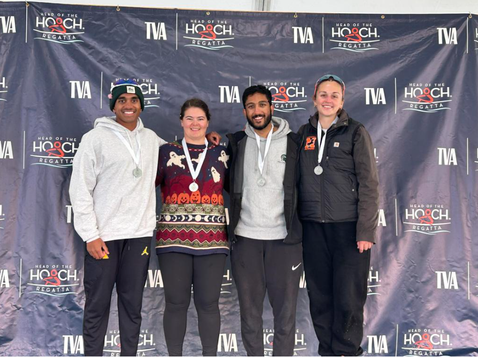 Group of four athletes standing together on a podium, each wearing medals around their necks, celebrating at the Head of the Hoach Regatta with a branded backdrop behind them.