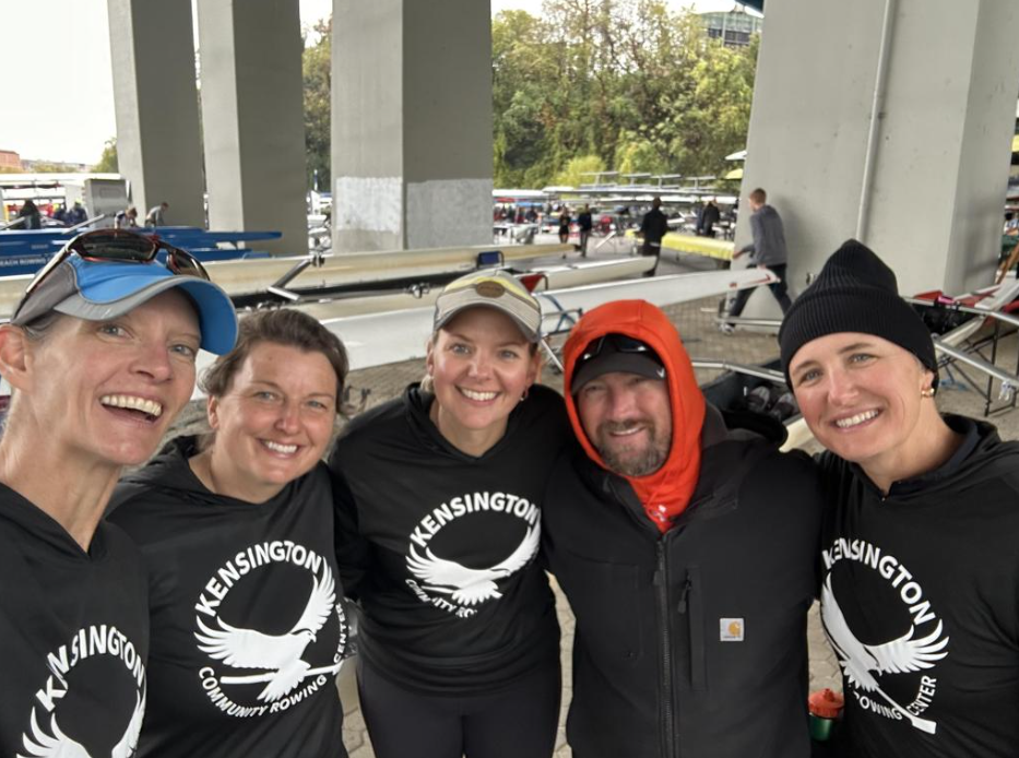 Group of five smiling people wearing black Kensington Community Rowing shirts, standing under a bridge, with rowing shells and other people in the background.