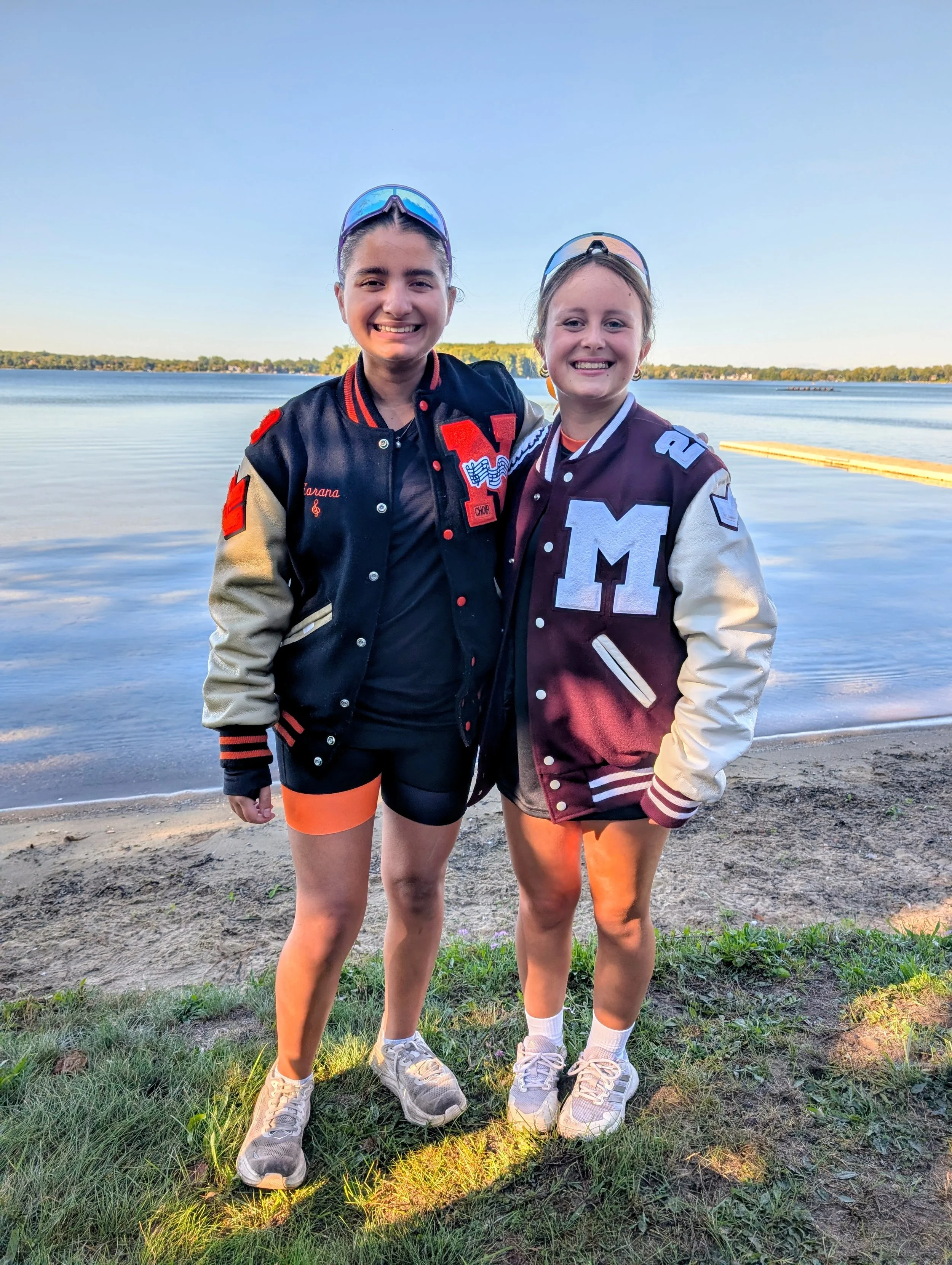 Two young women standing on a grassy shoreline by a lake, smiling at the camera, wearing varsity jackets, athletic shorts, sneakers, and sunglasses, with calm water and a small boat in the background during sunset.