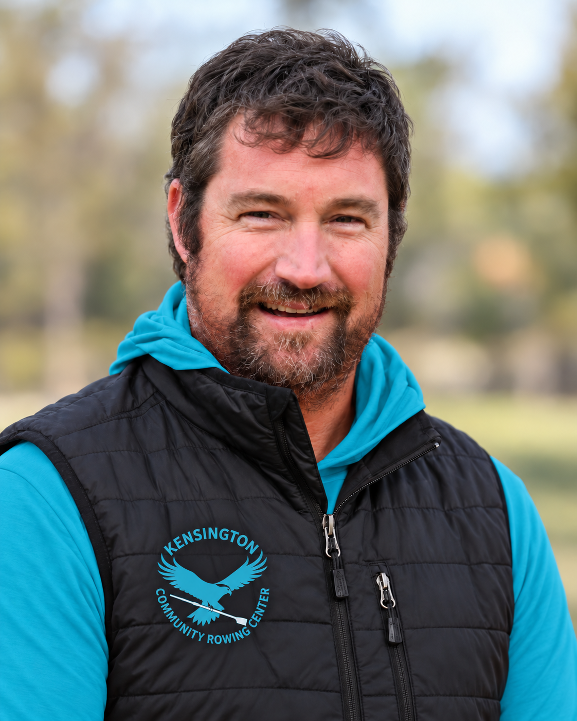 A man smiling outdoors, wearing a blue hoodie and a black vest with the emblem of Kensington Community Rowing Center.