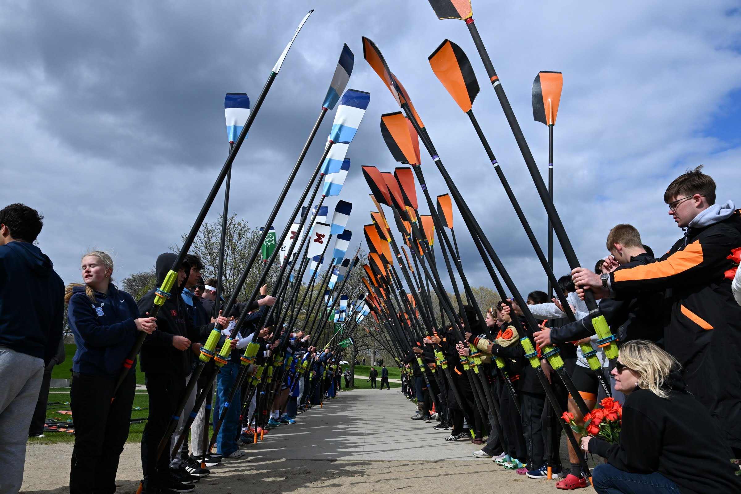 People standing in two rows holding paddles during a memorial or vigil, with some holding flowers, outdoors under cloudy sky.