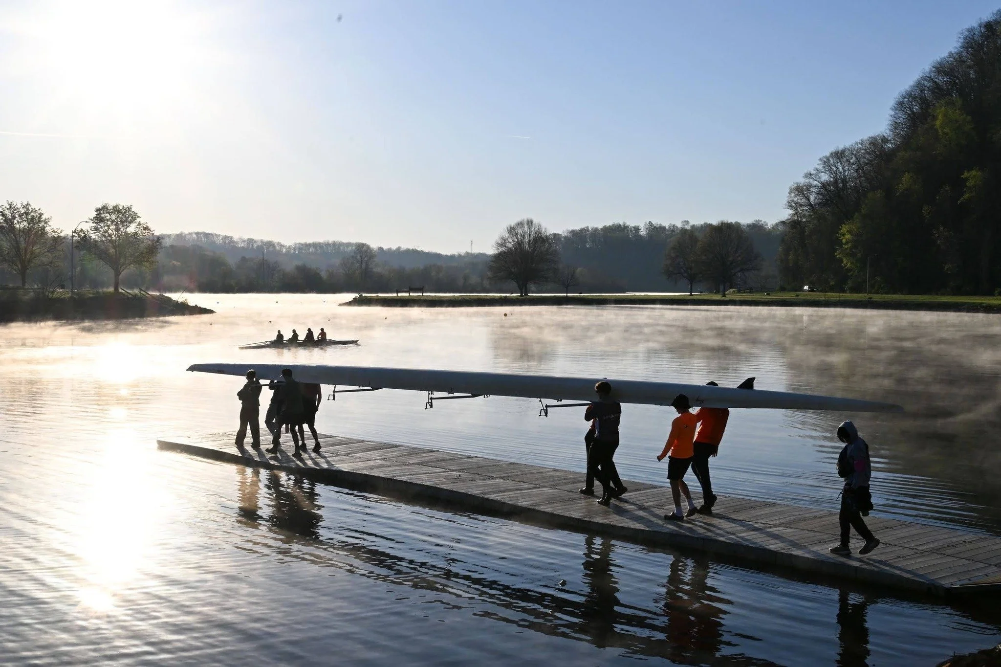 People preparing a boat on a dock by a calm river with trees along the shoreline and hills in the background under a bright sky.