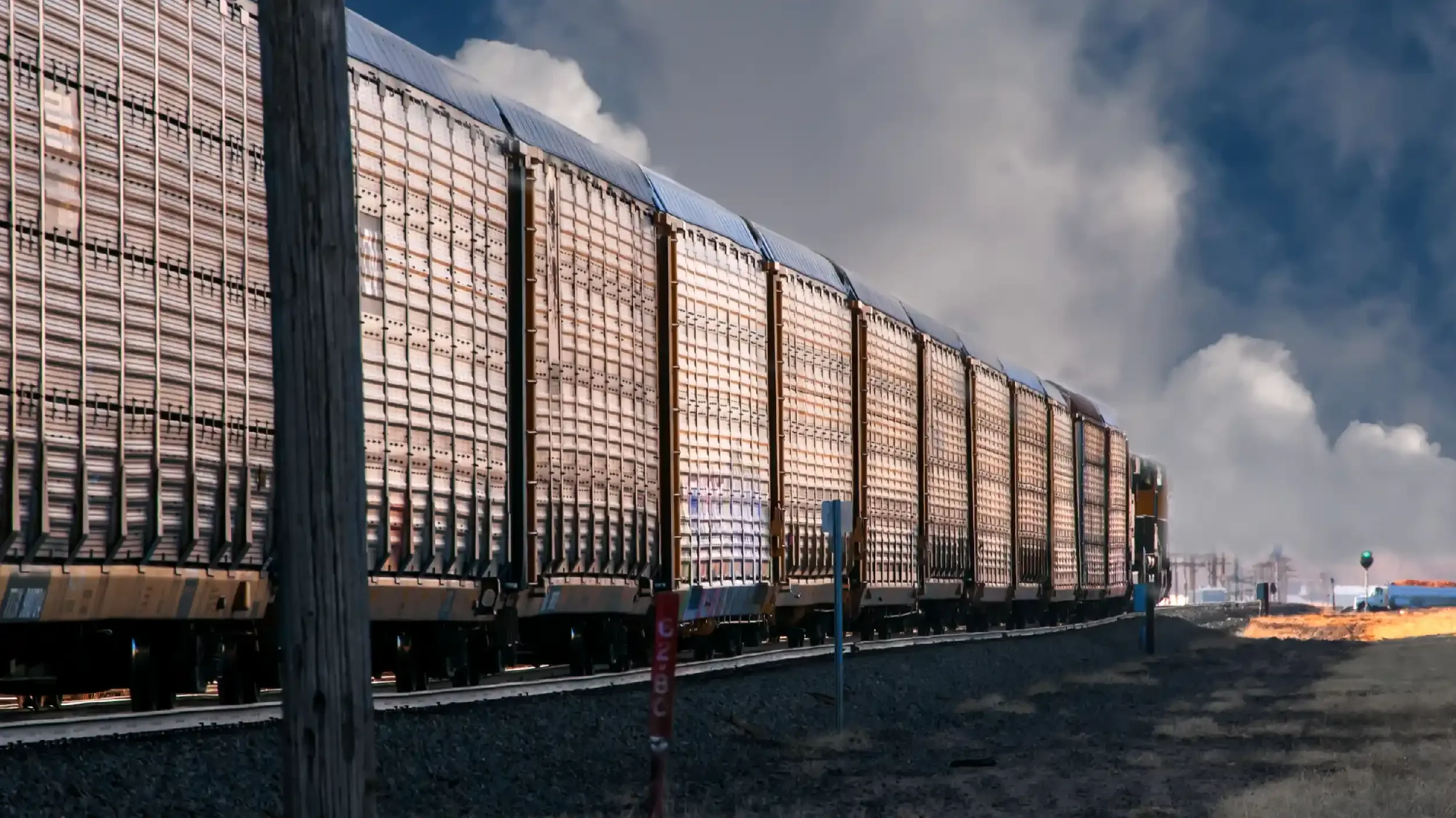 A long train with metal freight cars traveling on railway tracks with a clear sky and clouds in the background.