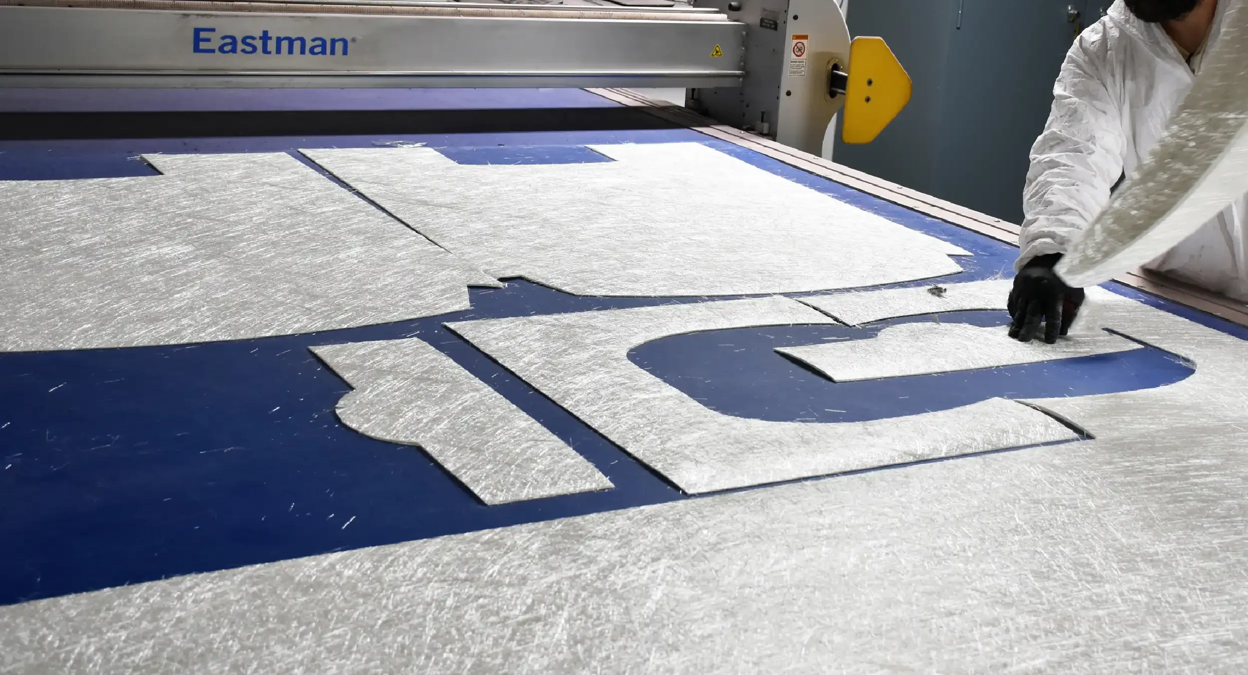 A worker arranges cut pieces of metallic foil on a blue surface, next to a large industrial machine labeled 'Eastman.'