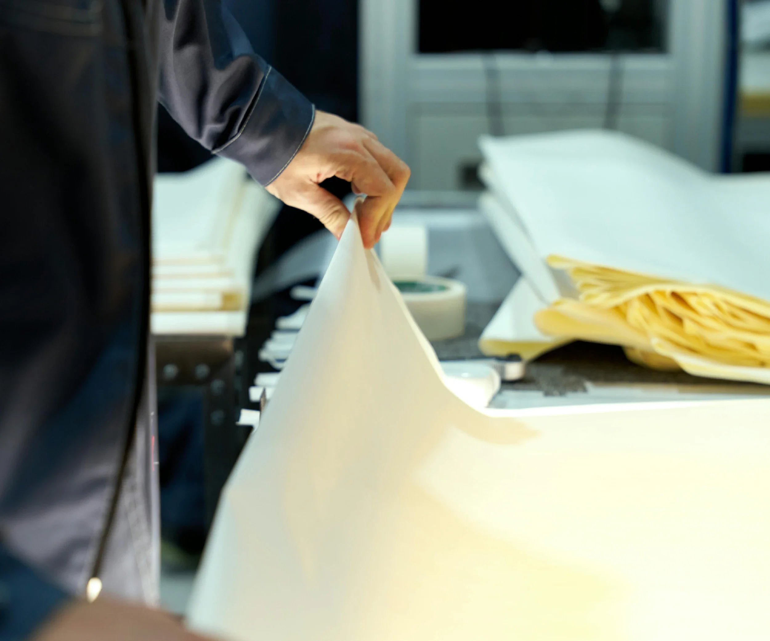 Close-up of a person's hand reaching into a container, with yellow fabric or paper on a table nearby, and a blurred background including a computer monitor and desk.