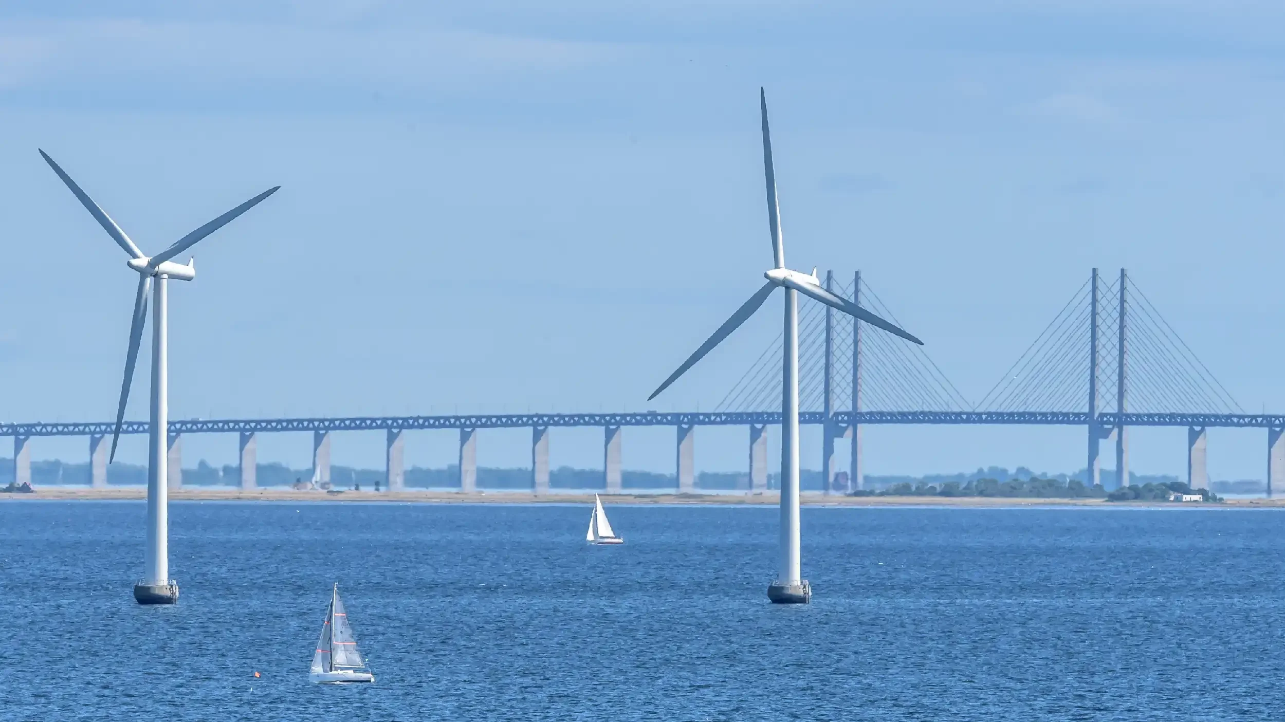 Wind turbines near a body of water with sailboats and a large bridge in the background.
