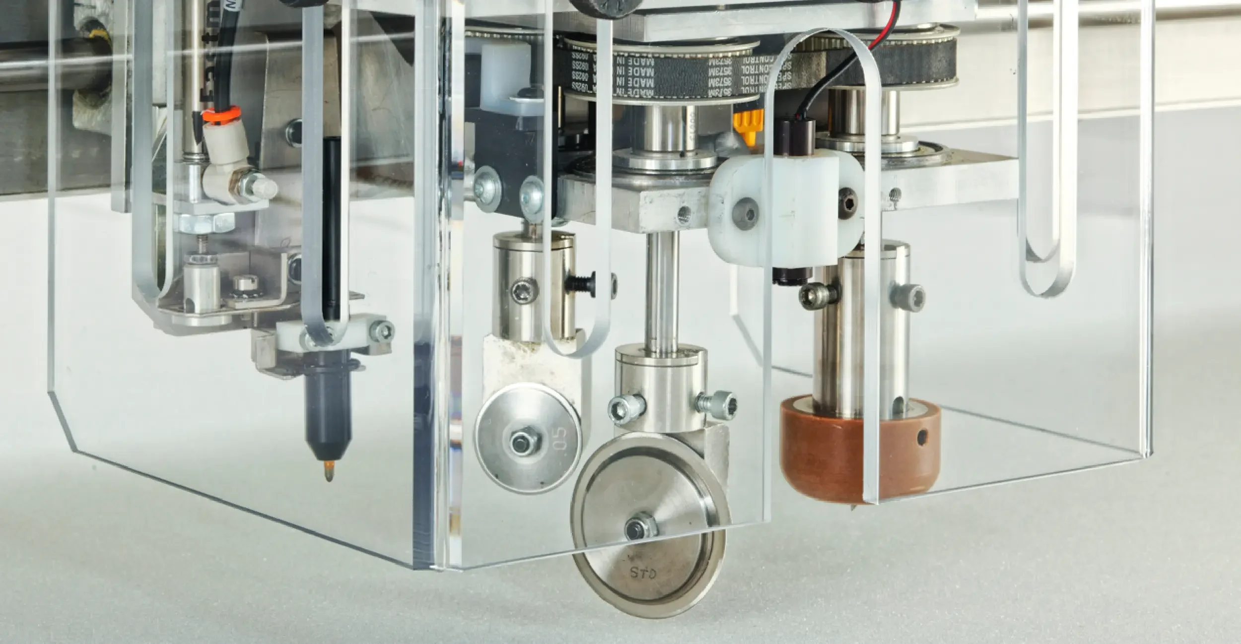 Close-up of a mechanical assembly with metal components, gears, belts, and wires inside a transparent enclosure.