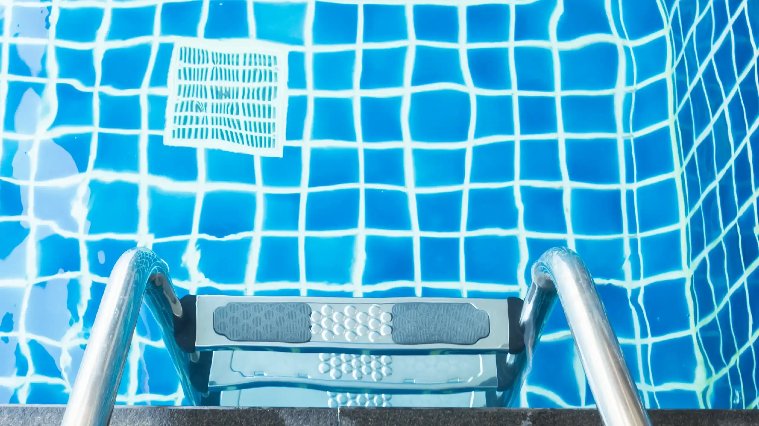View of a swimming pool from the perspective of someone standing on a step ladder. The pool has a blue tiled floor with white grout lines and a metal handrail at the pool's edge. A gray non-slip step platform with textured patterns is in the foreground.