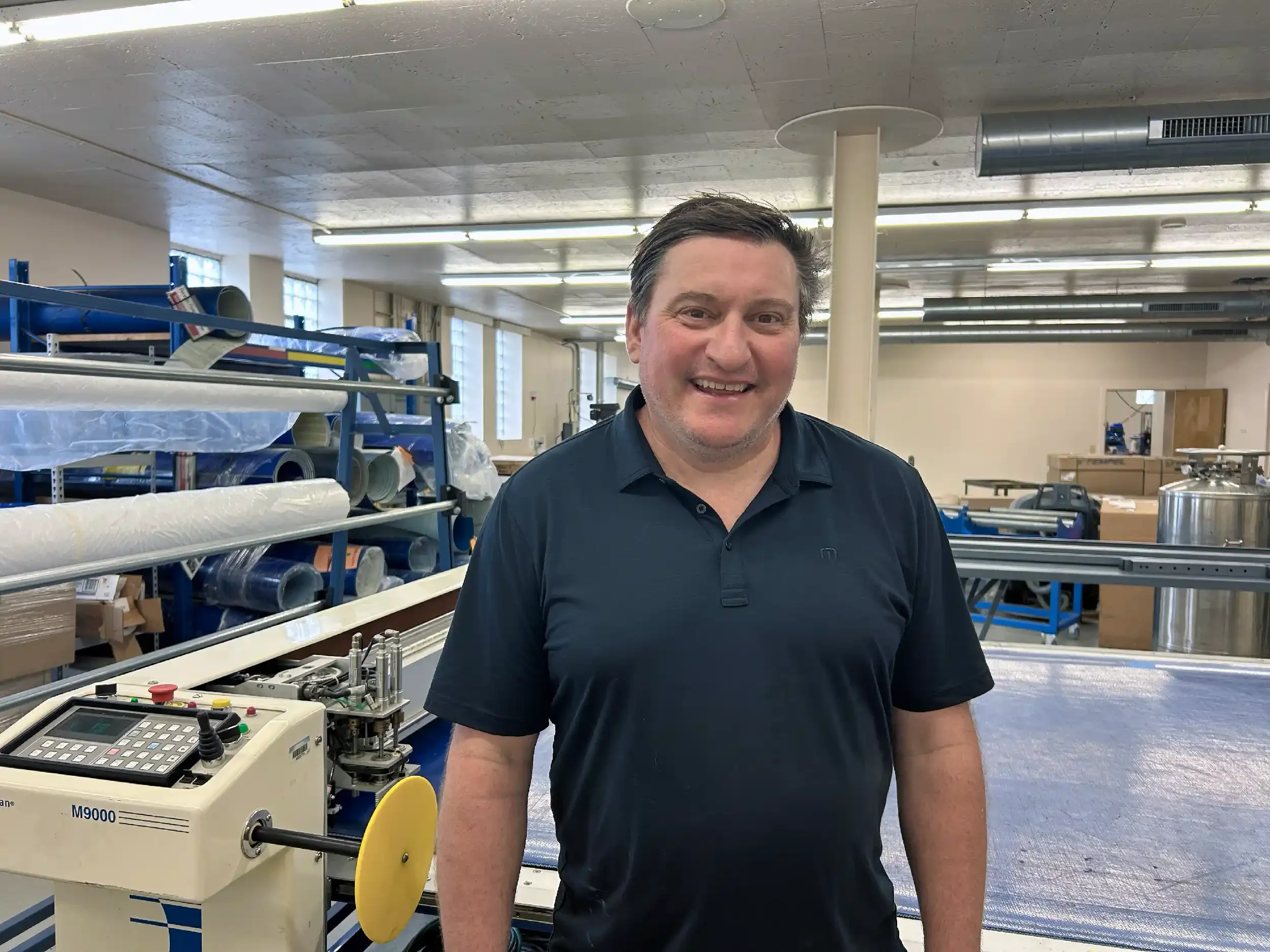 A smiling man with dark hair and a navy blue polo shirt standing in a manufacturing space, surrounded by industrial cutting equipment, shelves with rolls of material, and machinery.