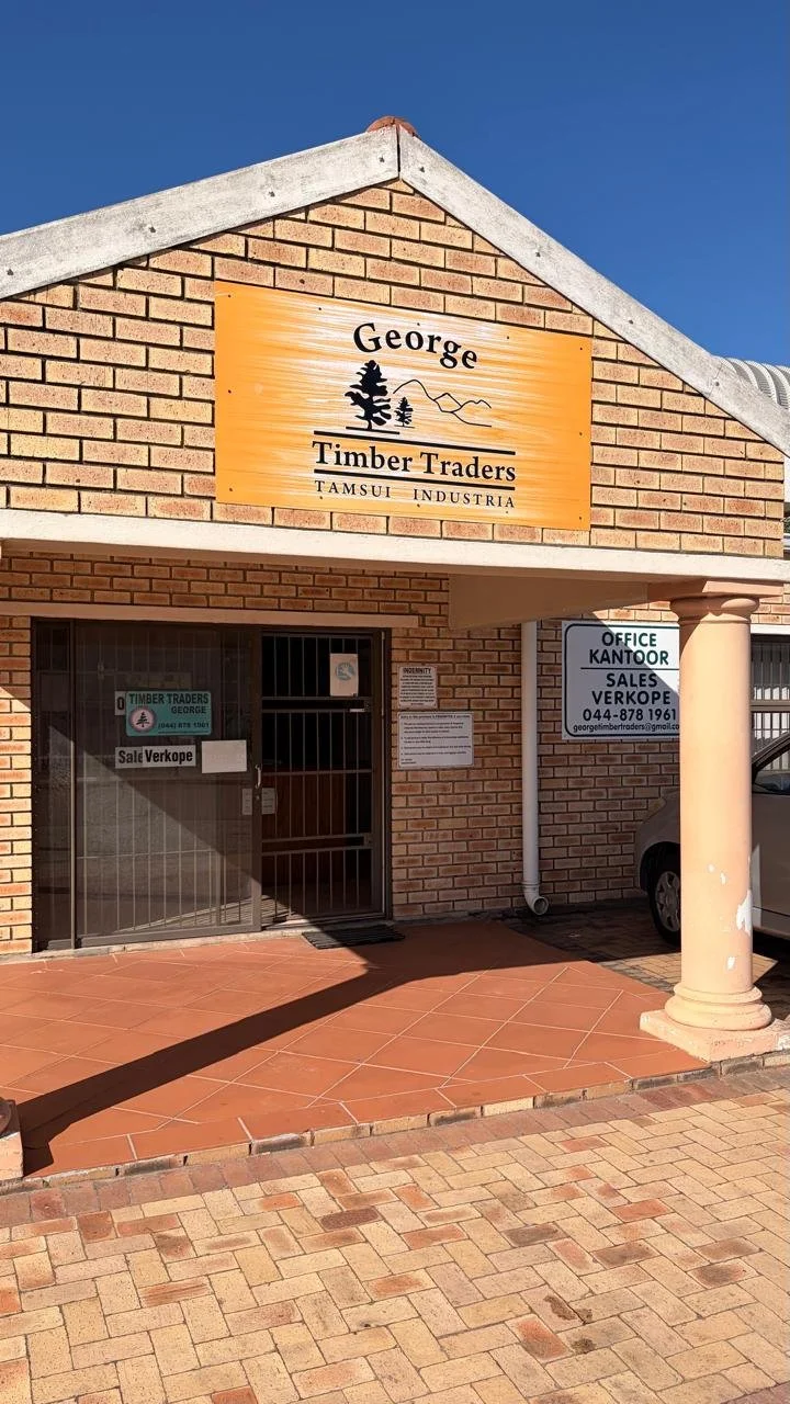A brick building with a sign that reads "George Timber Traders." The sign features mountain and tree illustrations. There are signs on the door and wall advertising timber trading and sales, with contact details. A column supports the porch area, and part of a parked car is visible on the right side.