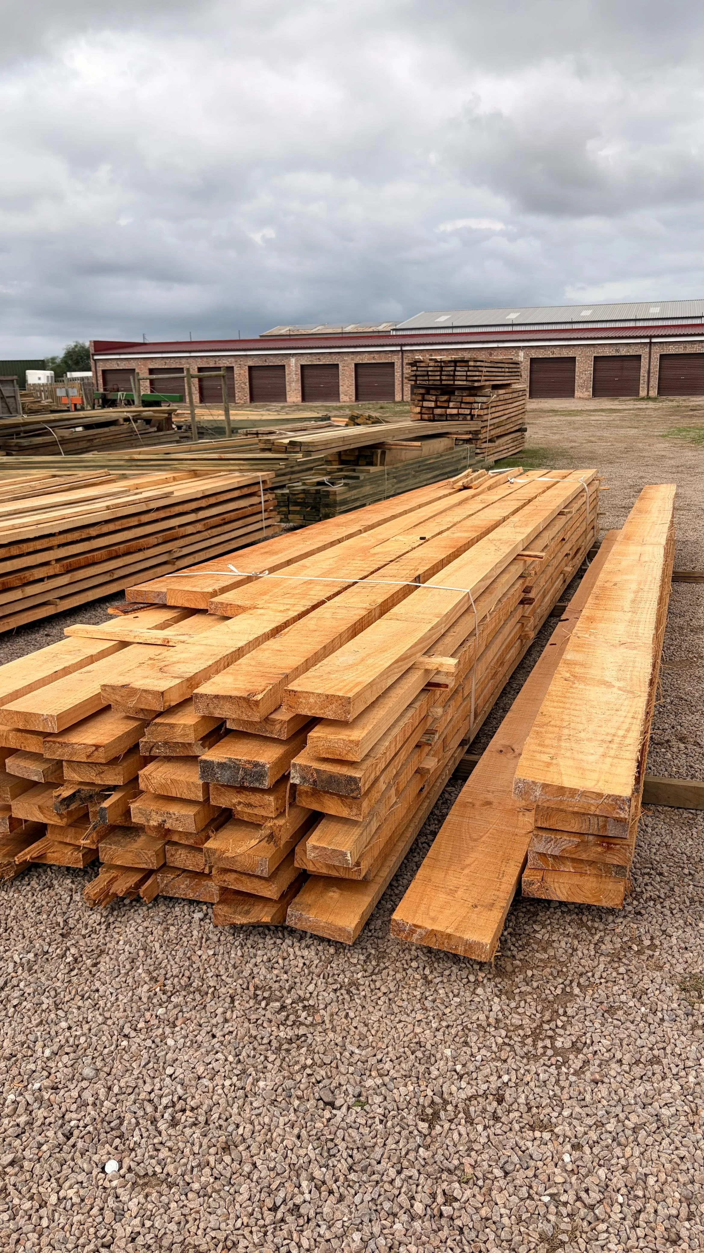 Stacks of wooden planks and lumber on gravel outside, with a brick building with multiple garage doors in the background and a cloudy sky overhead.