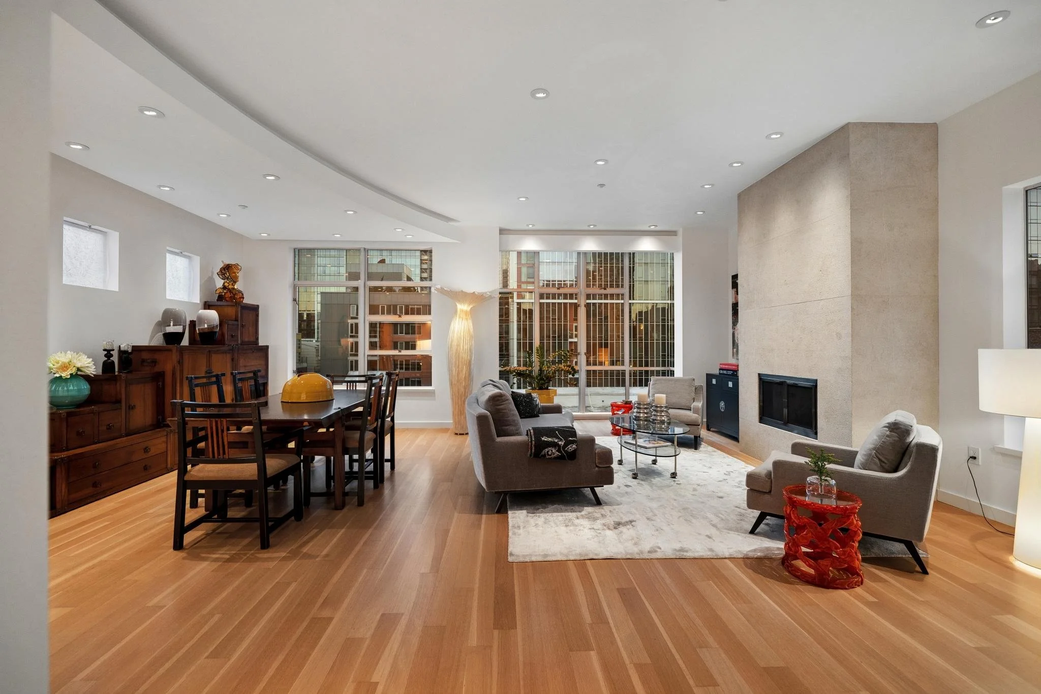 Interior of a modern living room with wooden flooring, gray and white furniture, large windows showing city buildings, and various decorative items.