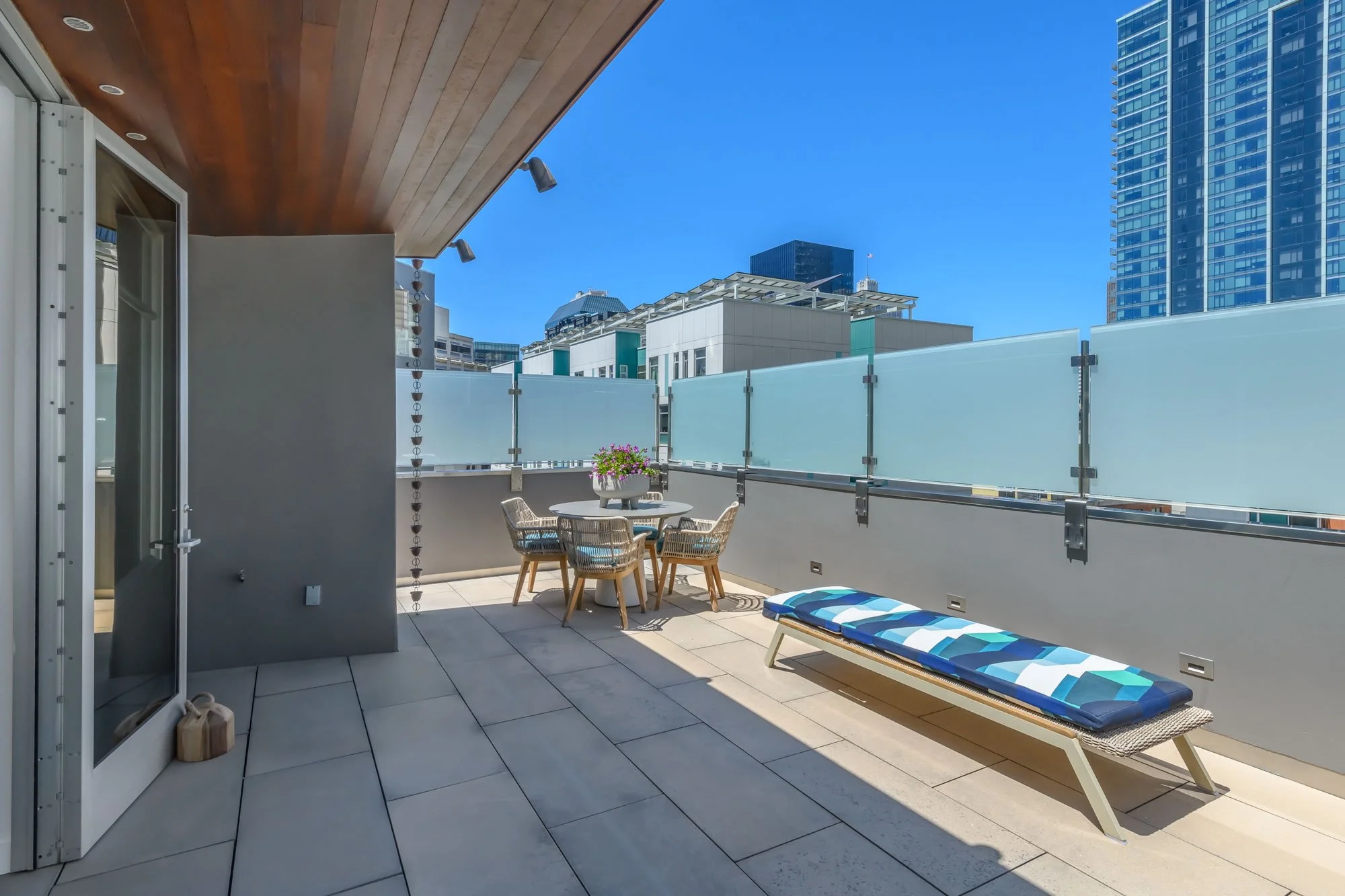 City balcony with outdoor furniture, planter with pink flowers, lounge chair with blue and white striped cushion, high-rise buildings in the background, clear blue sky.