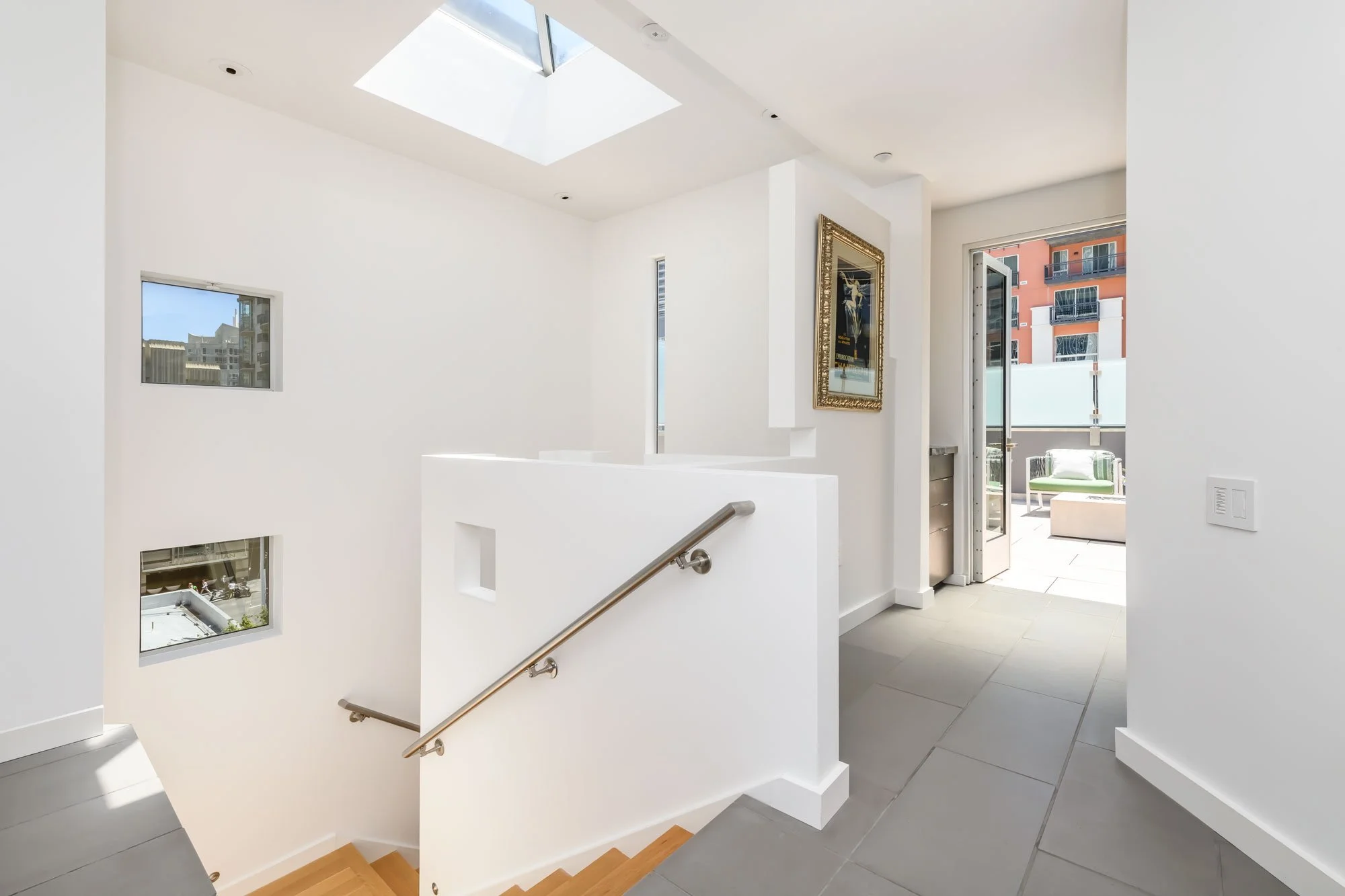 Interior view of a modern, white-painted apartment staircase with multiple windows allowing natural light, a framed artwork on the wall, and a balcony with outdoor furniture visible through an open door.