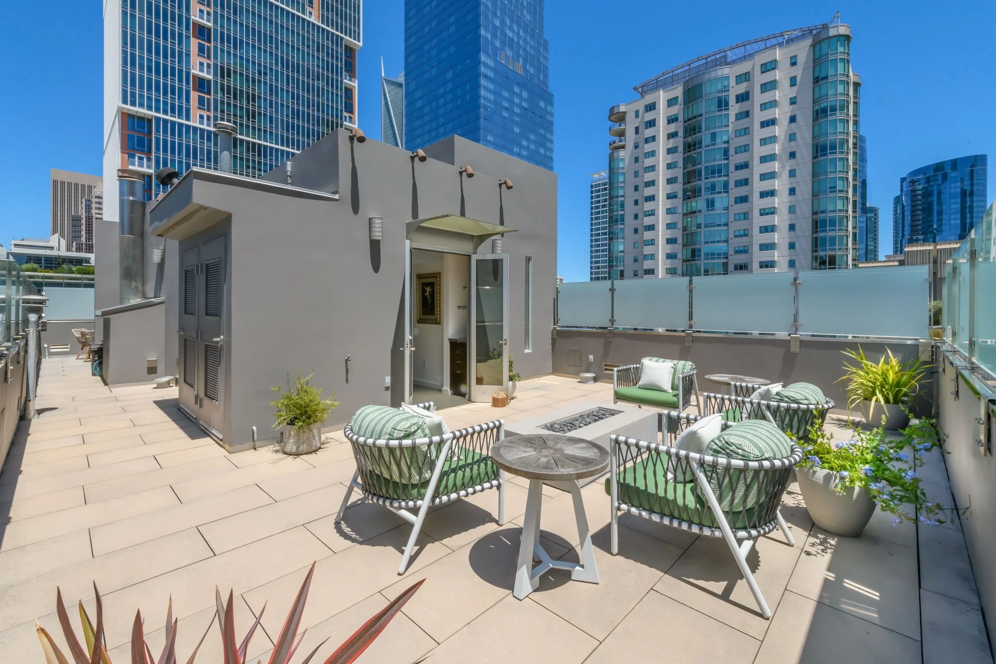 Rooftop patio with outdoor seating including chairs and a small table, surrounded by glass railings, with tall modern skyscrapers in the background against a clear blue sky.