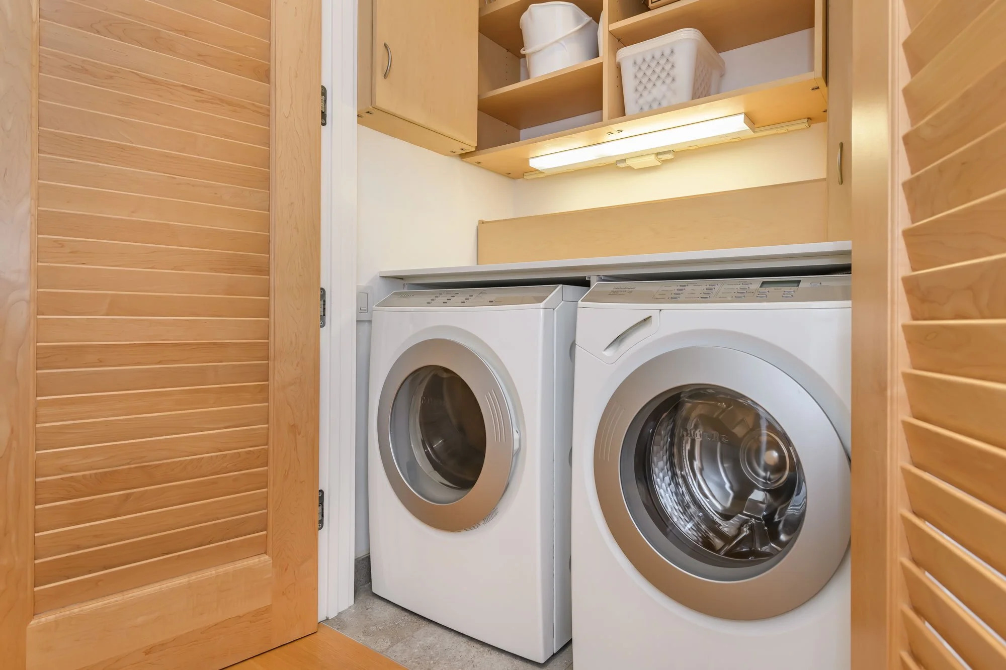 A laundry area inside a closet with wooden sliding doors, featuring a washing machine and dryer side by side. The top shelf holds laundry baskets and a container, and a fluorescent light illuminates the space.