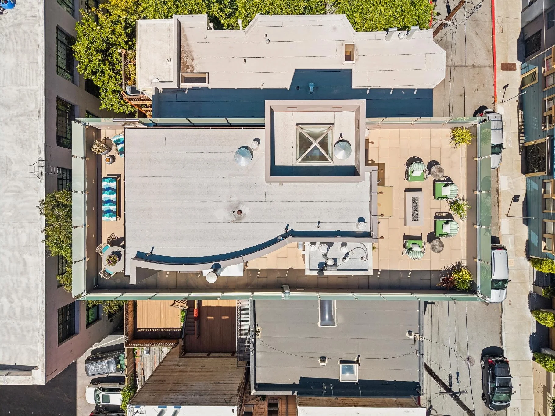 An aerial view of a building rooftop with a small outdoor seating area, planters, and chairs. The rooftop is divided into sections with different flooring materials, including beige tiles and concrete, and features an architectural structure with a g