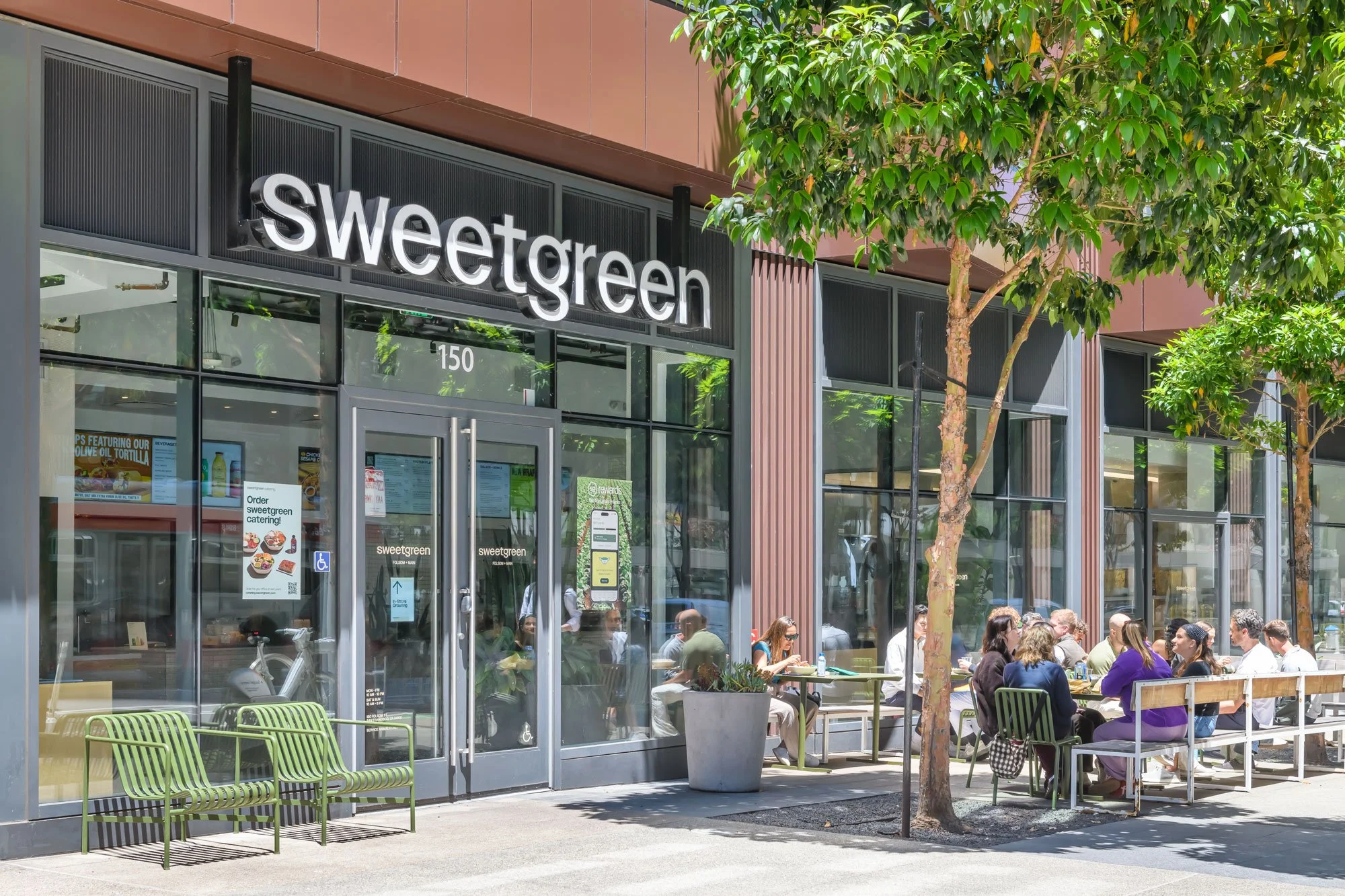Side view of a Sweetgreen restaurant with outdoor seating, people dining outdoors, and lush green trees.