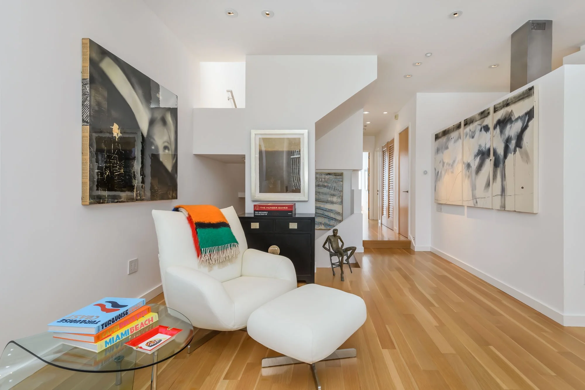 Living room with white walls and hardwood floors. Features modern white armchair with colorful striped blanket, glass coffee table with books, abstract wall art, and decorative sculpture. Natural light from a skylight and hallway lead to other areas.