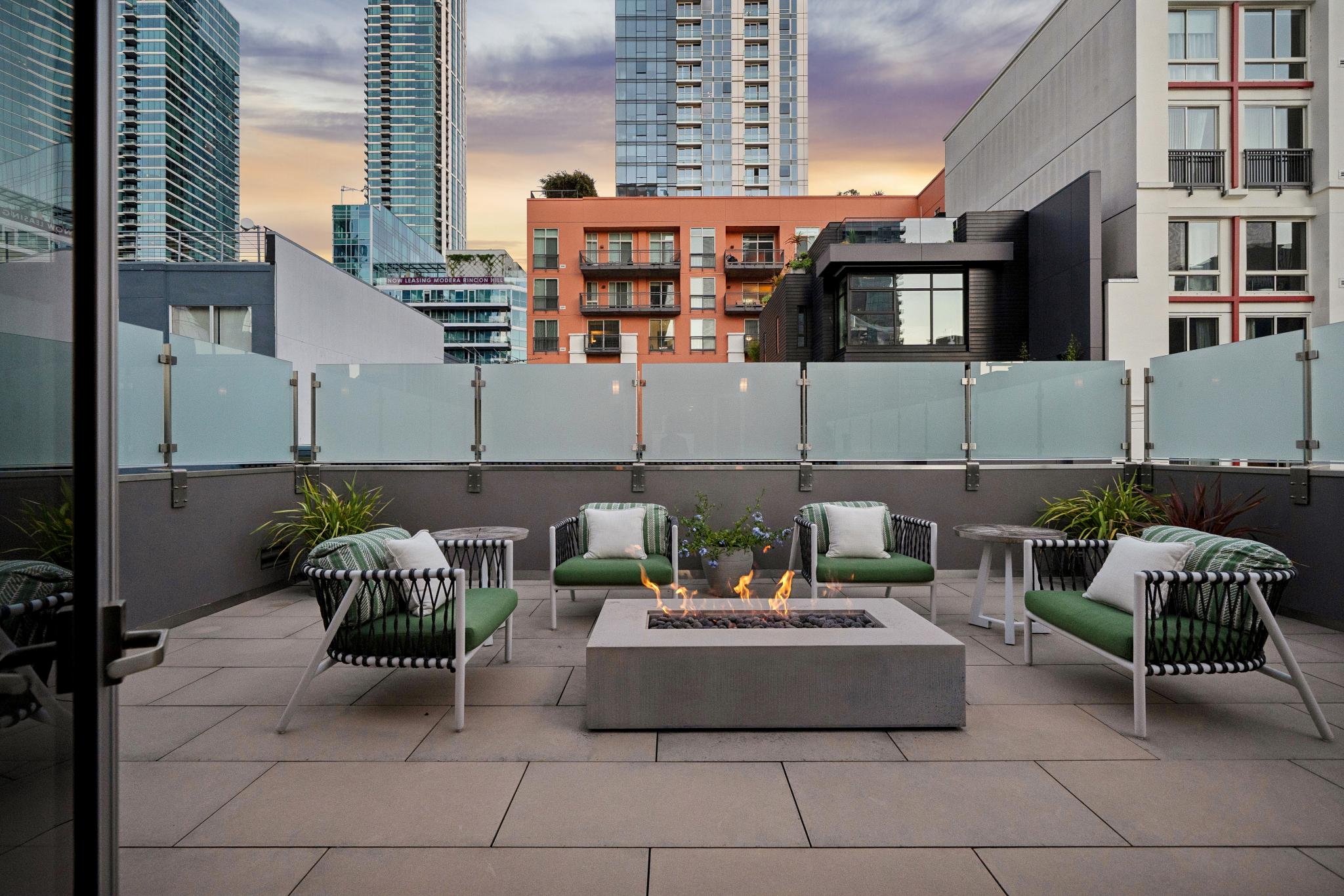 City rooftop patio with four green-cushioned chairs around a rectangular fire pit, potted plants, and a modern glass railing, overlooking tall urban buildings at sunset.