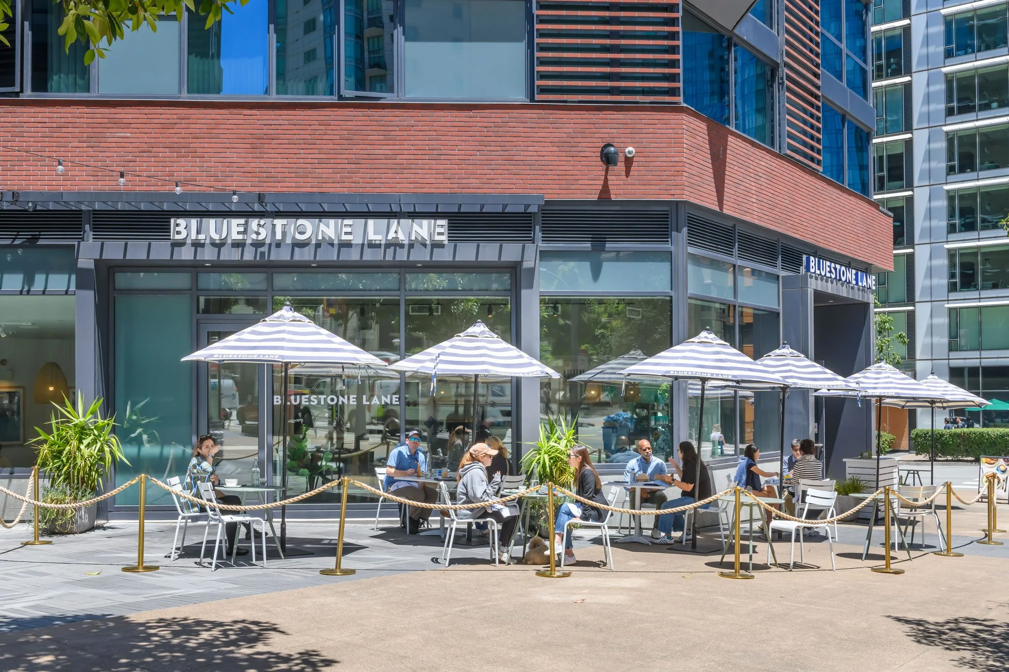 Outdoor patio of a cafe or restaurant with people sitting at tables under striped umbrellas, in front of a modern building with large glass windows and a sign that reads 'Bluestone Lane.'
