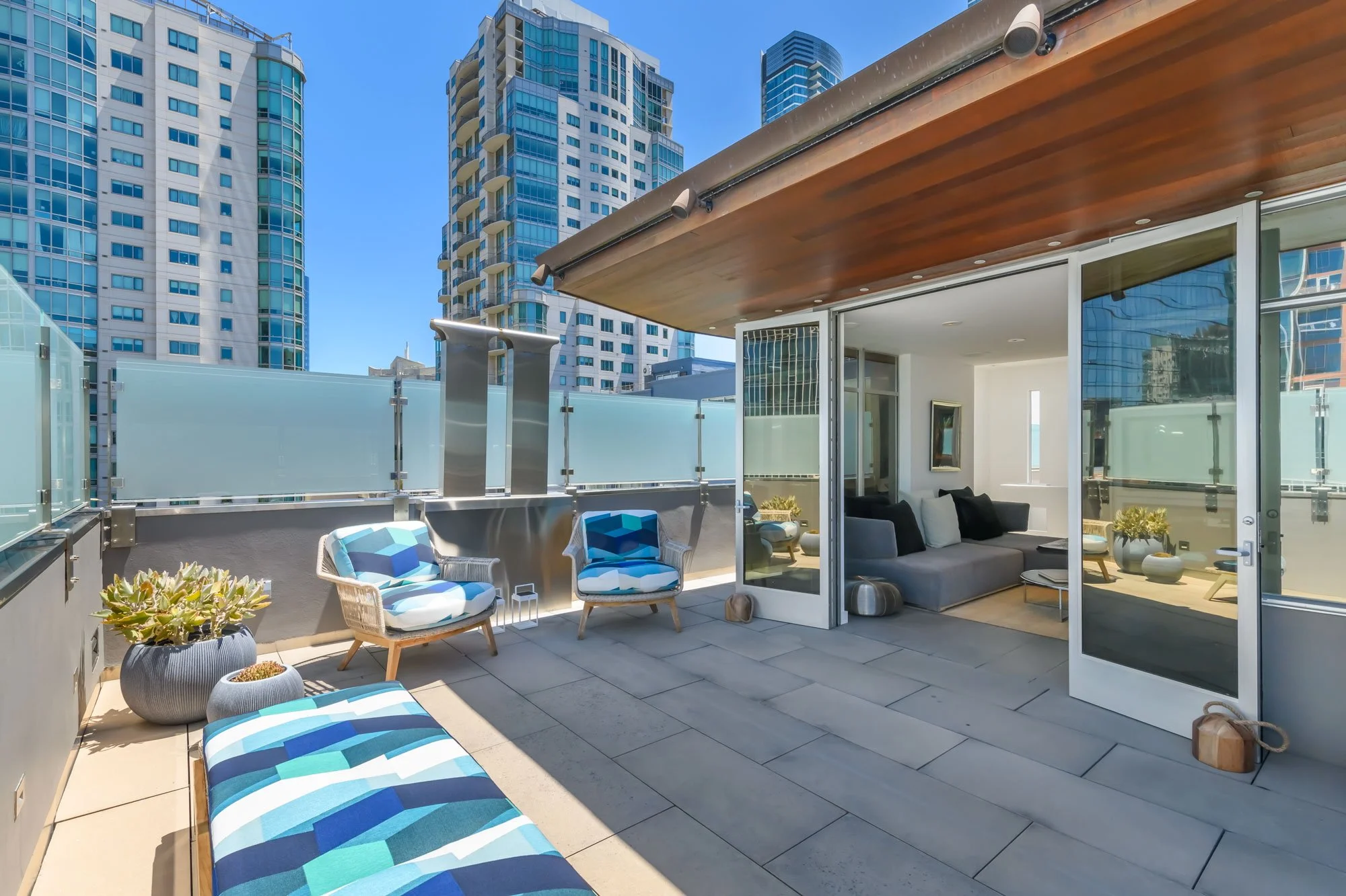 A modern rooftop patio with outdoor seating, including lounge chairs with blue and white striped cushions, a gray sofa, and potted plants, surrounded by glass railings with a city skyline of tall buildings in the background on a sunny day.
