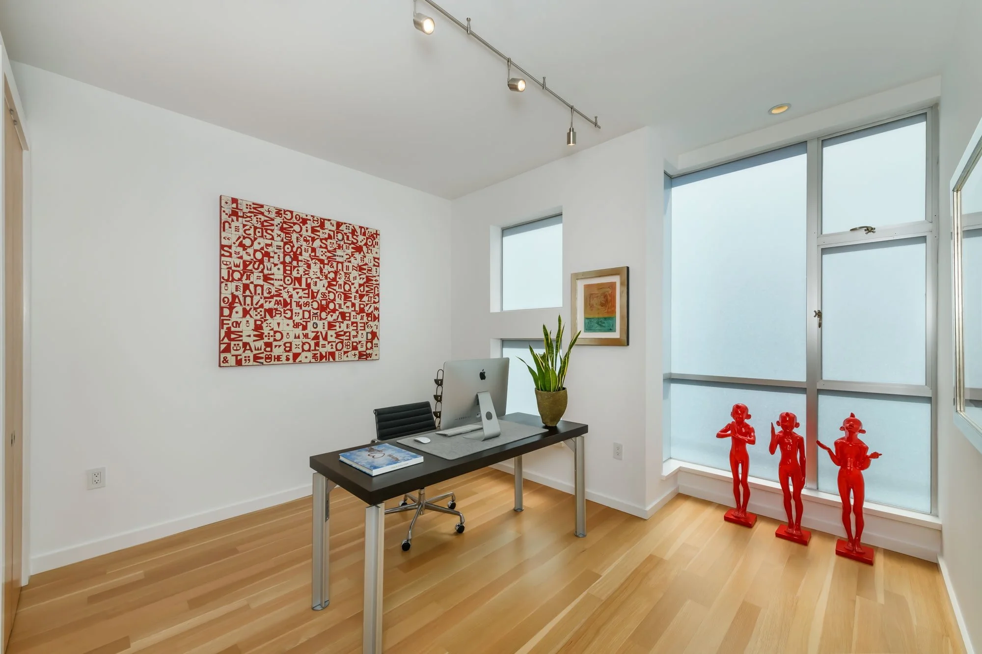 An office with white walls, hardwood floor, and large windows. There is a black desk with a computer, a plant, and magazines. Artwork decorates the walls, and red sculptures of children stand near the window.
