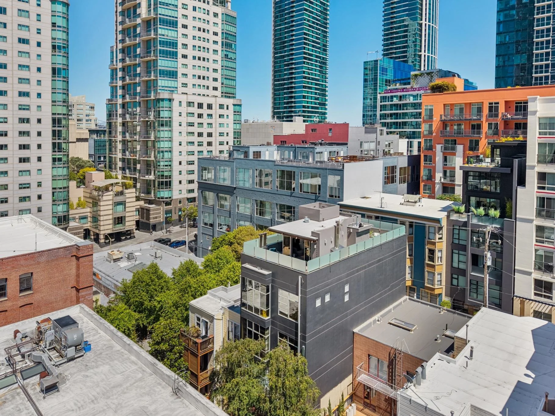 A cityscape featuring tall modern buildings, some with glass facades, and shorter older buildings with rooftops. Trees and streets are visible among the buildings, with a clear blue sky overhead.