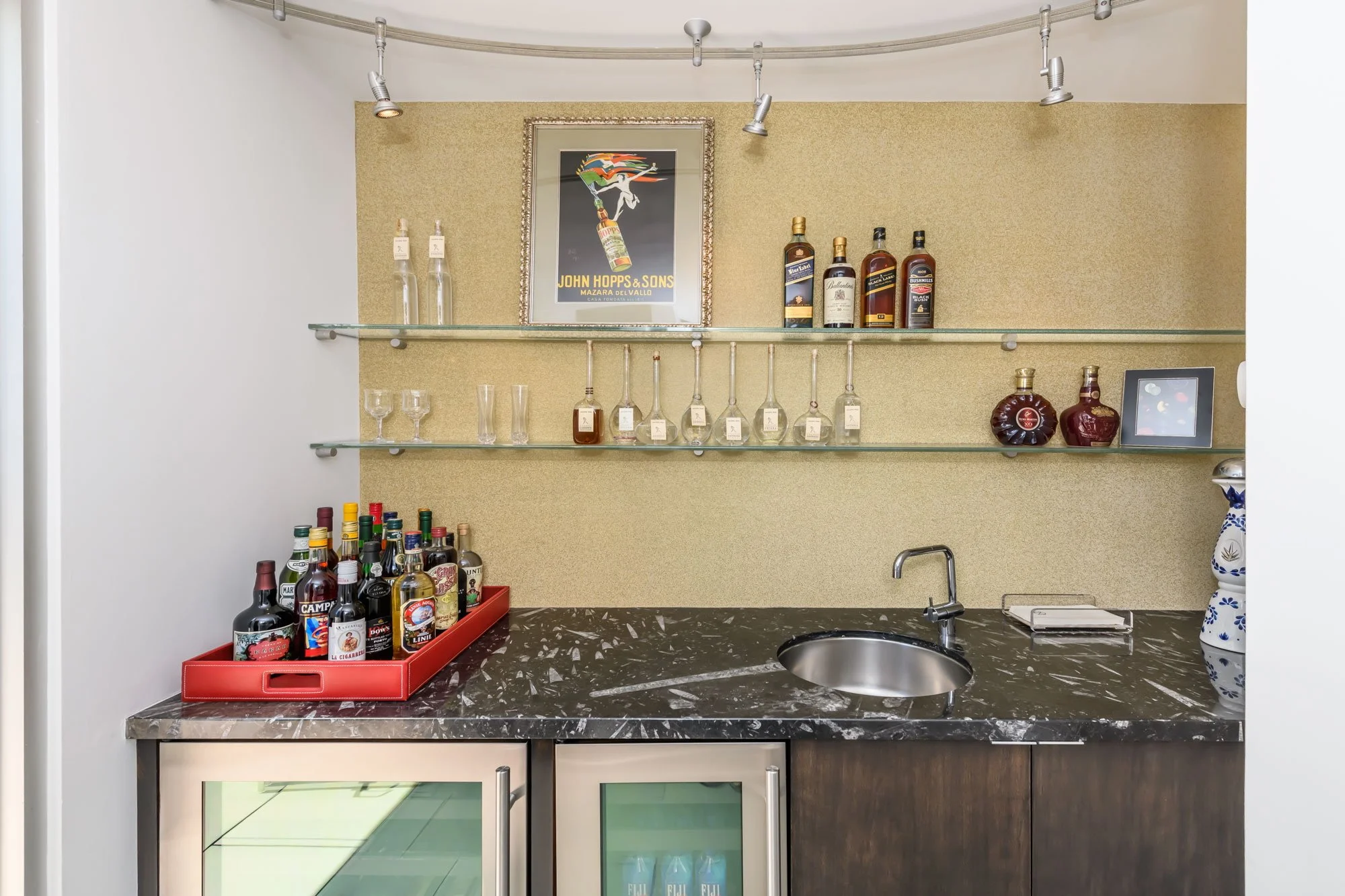 A kitchen bar area with glass shelves holding various liquor bottles, wine glasses, and a framed vintage advertisement poster. The countertop has a tray of liquor bottles, and a decorative blue and white vase is on the right side. The wall has a text