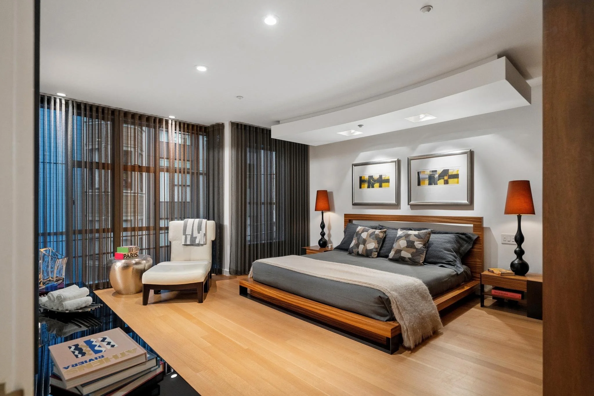 Modern bedroom with wooden bed frame, gray bedding, two patterned pillows, two black bedside lamps, large artwork above bed, white wall, and wooden side table. Large windows with sheer and brown curtains allow natural light. Light wood flooring.
