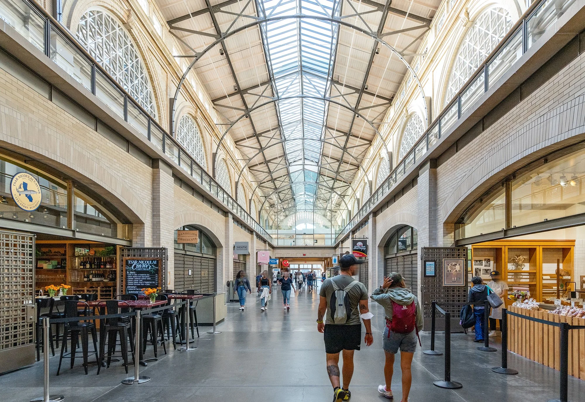 Indoor shopping mall with shoppers walking, stores on either side, high arched glass ceiling, and outdoor cafe seating.