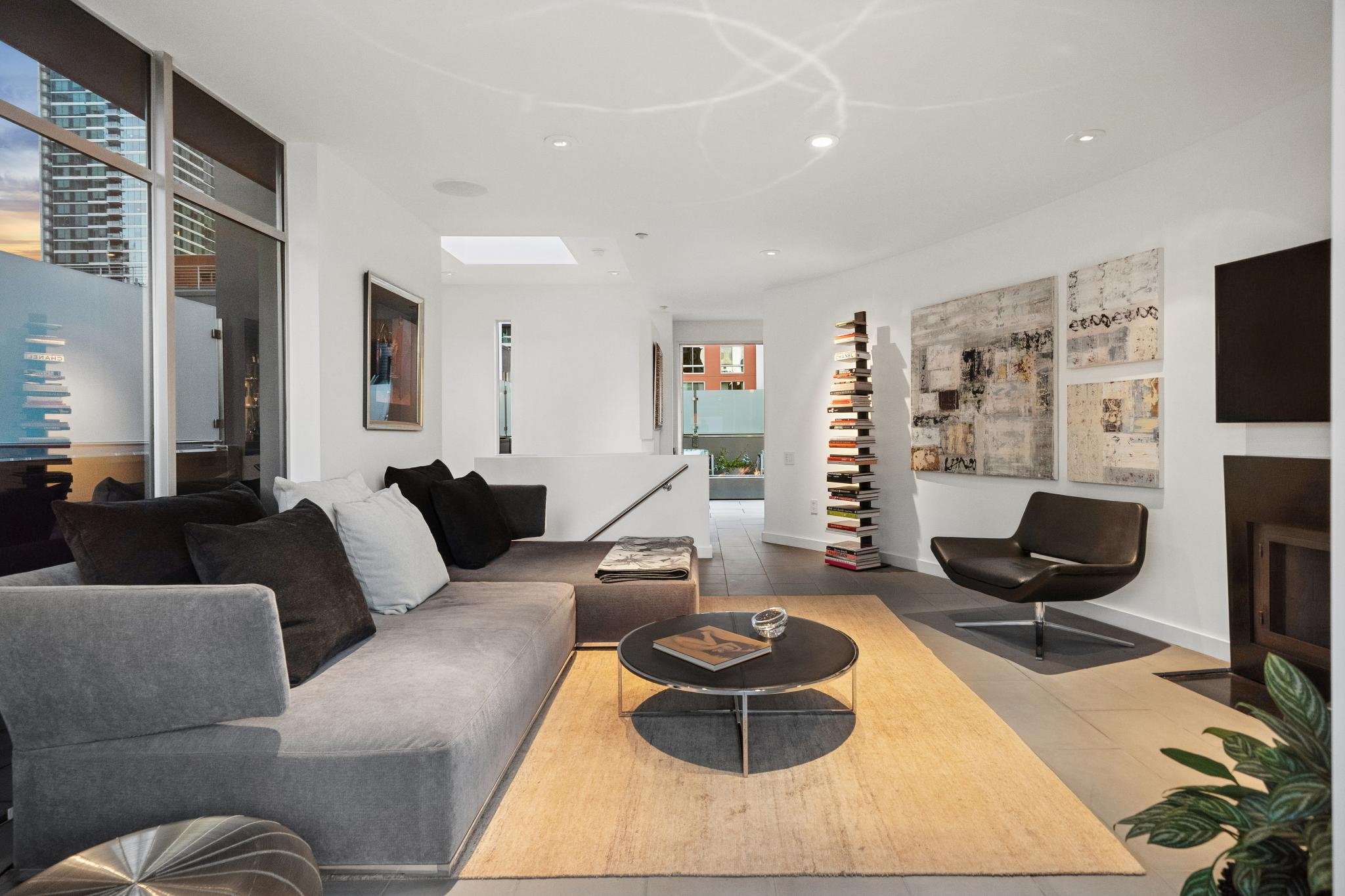 A modern living room with a gray sectional sofa with black and white pillows, a round coffee table with books, abstract wall art, a black lounge chair, and a tall stack of books. Large windows show city buildings outside, and the room has a white cei