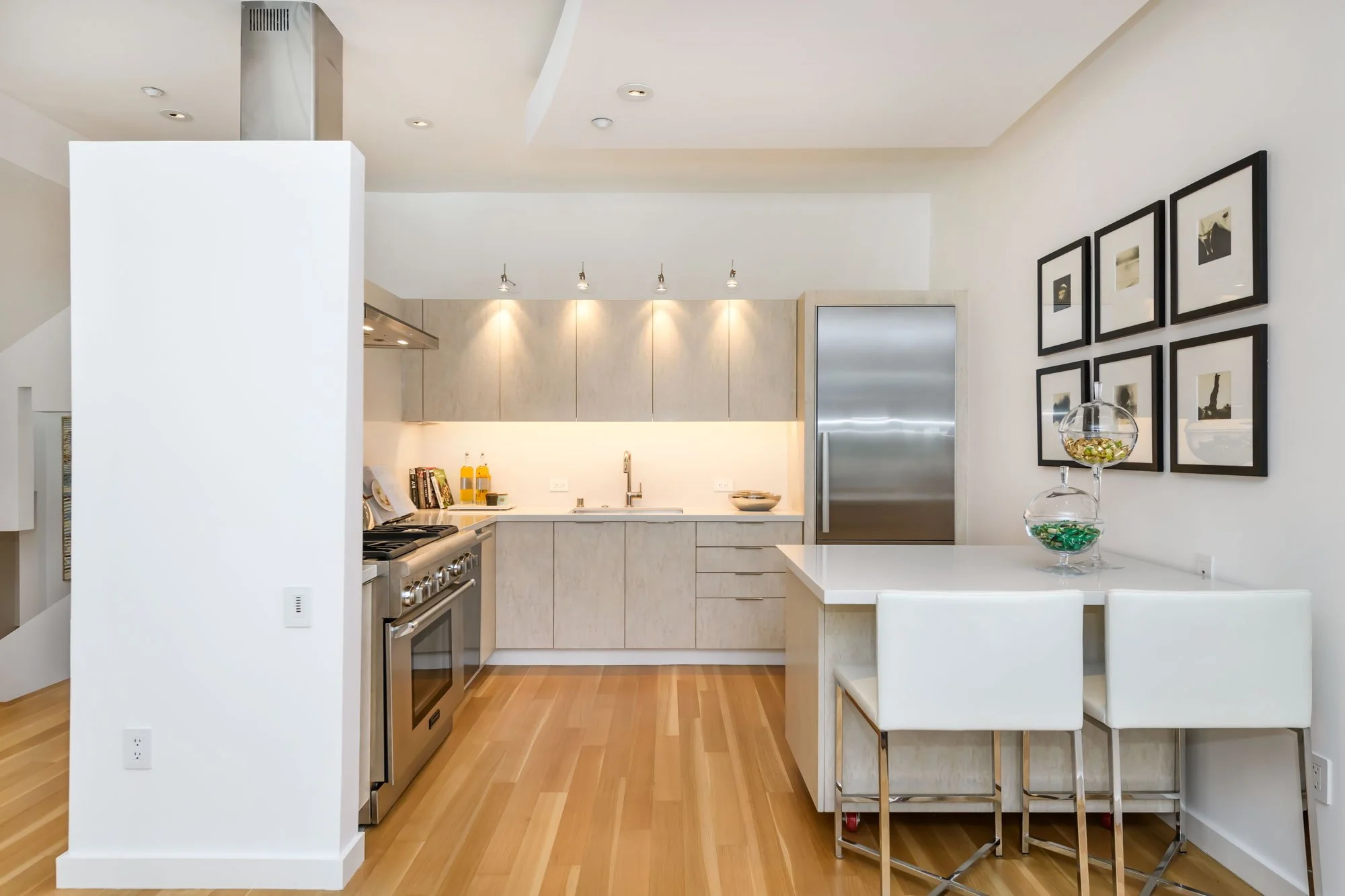 Modern kitchen with white cabinetry, stainless steel appliances, framed black-and-white art on the wall, glass bowls with decorative items on the counter, and light wood flooring.