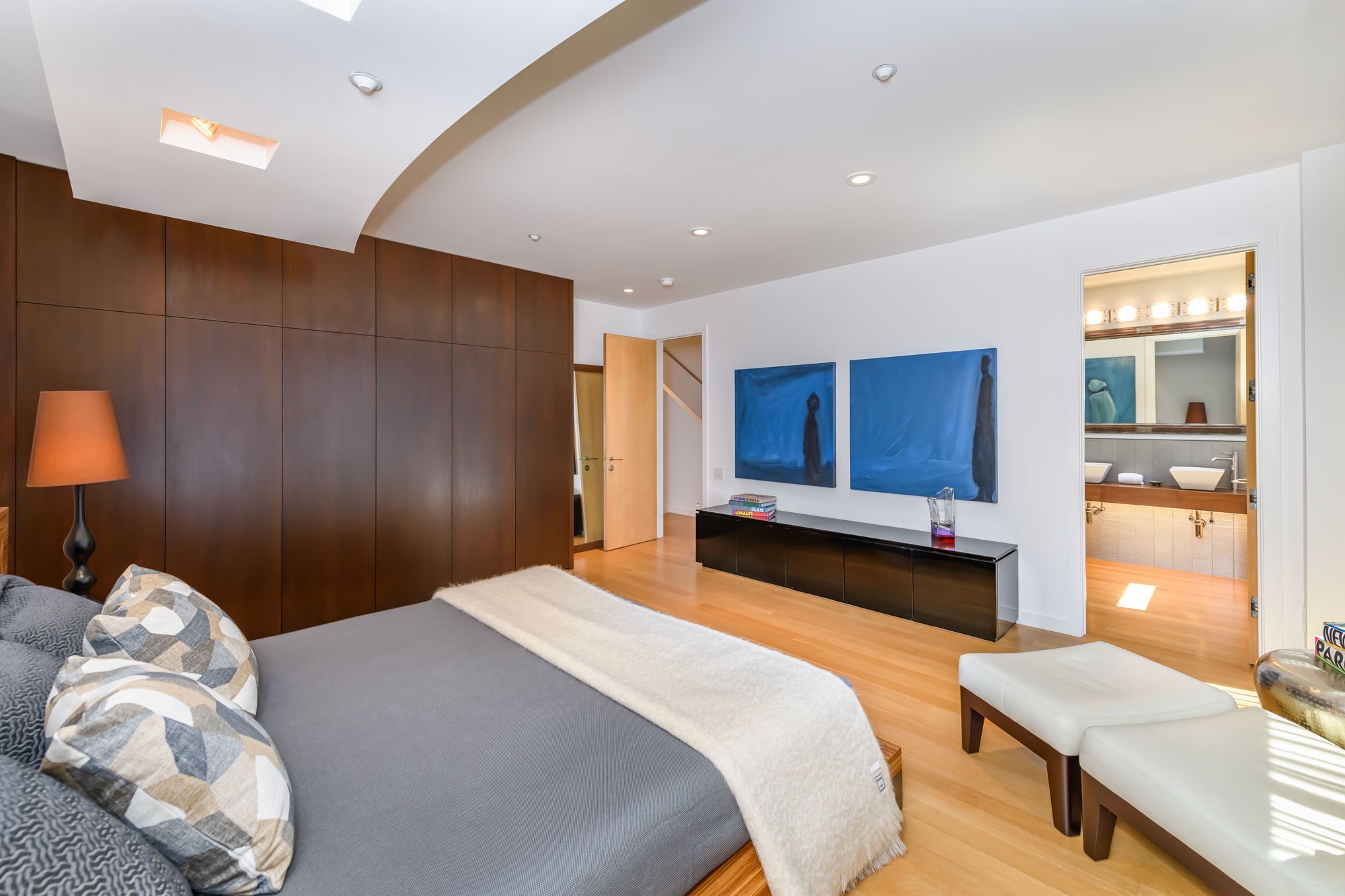 Modern living room with white walls, wooden floor, a dark cabinet, two blue artworks, and a bathroom visible through an open door.