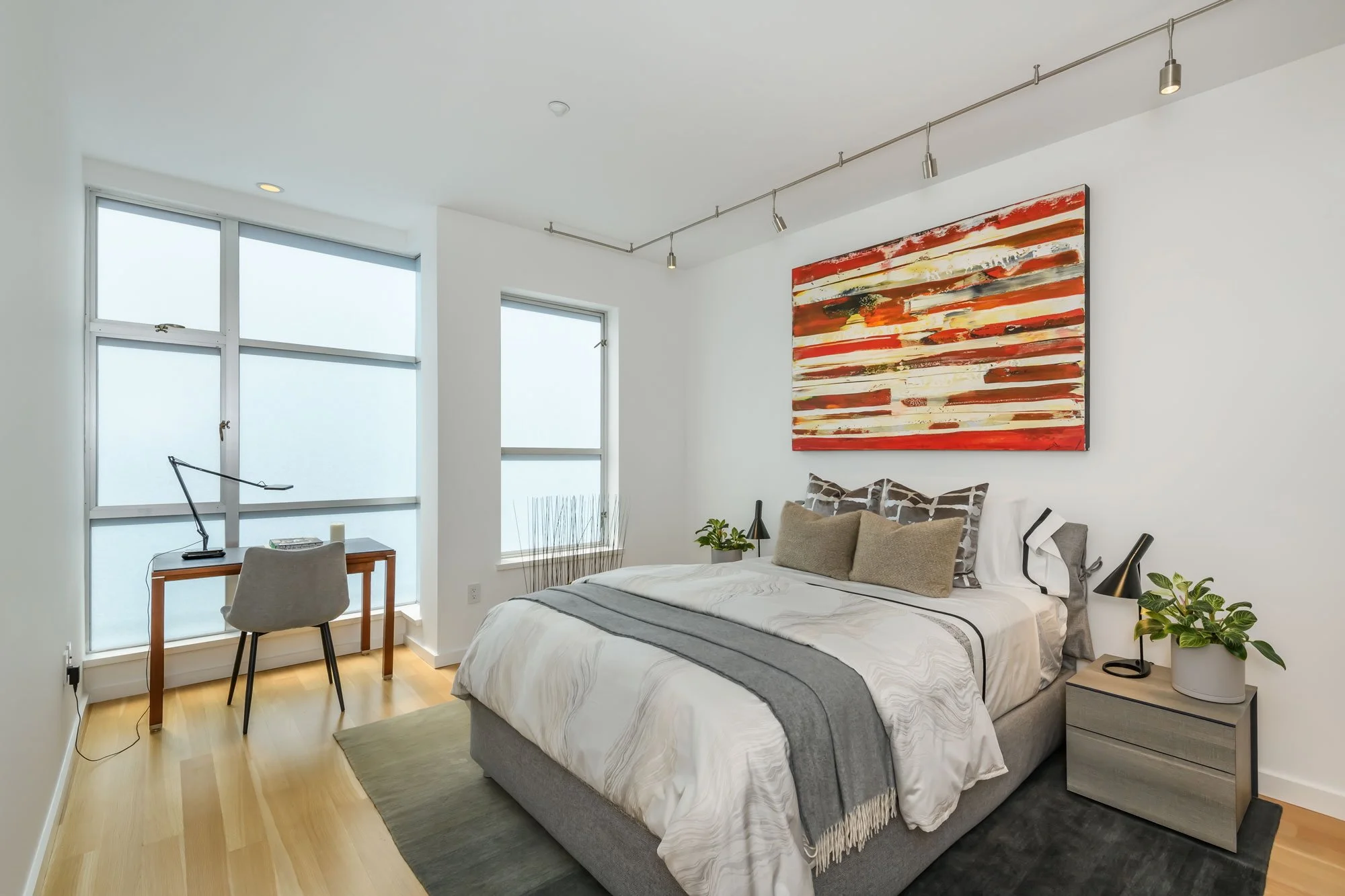 Modern bedroom with large windows, abstract painting on the wall, bed with beige pillows, bedside table with green plant, desk with lamp, and wooden floor.