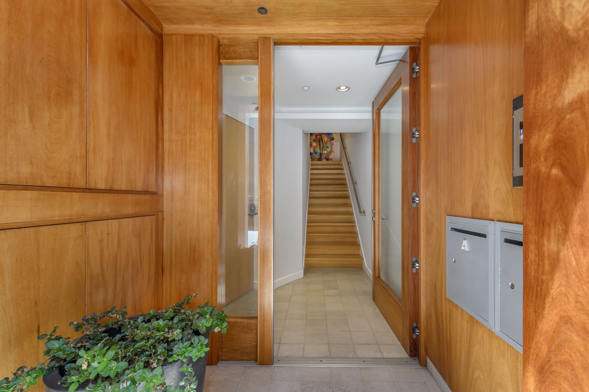 A wooden reception desk with a small potted plant and an elevator control panel, leading to a staircase in a hallway with white walls and beige tiled flooring.