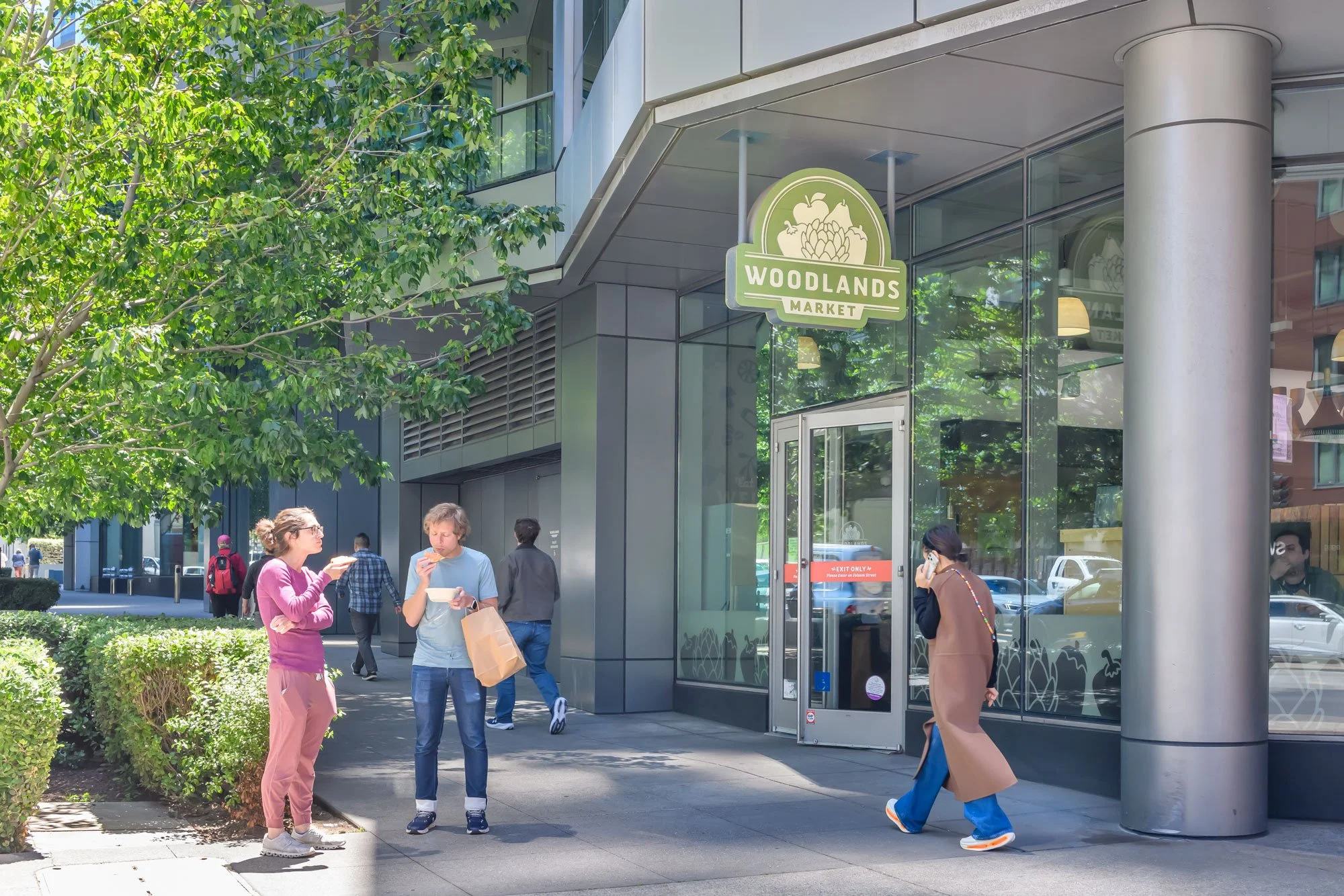 People walking and eating outside of Woodland Markets storefront, with green trees and modern building architecture.