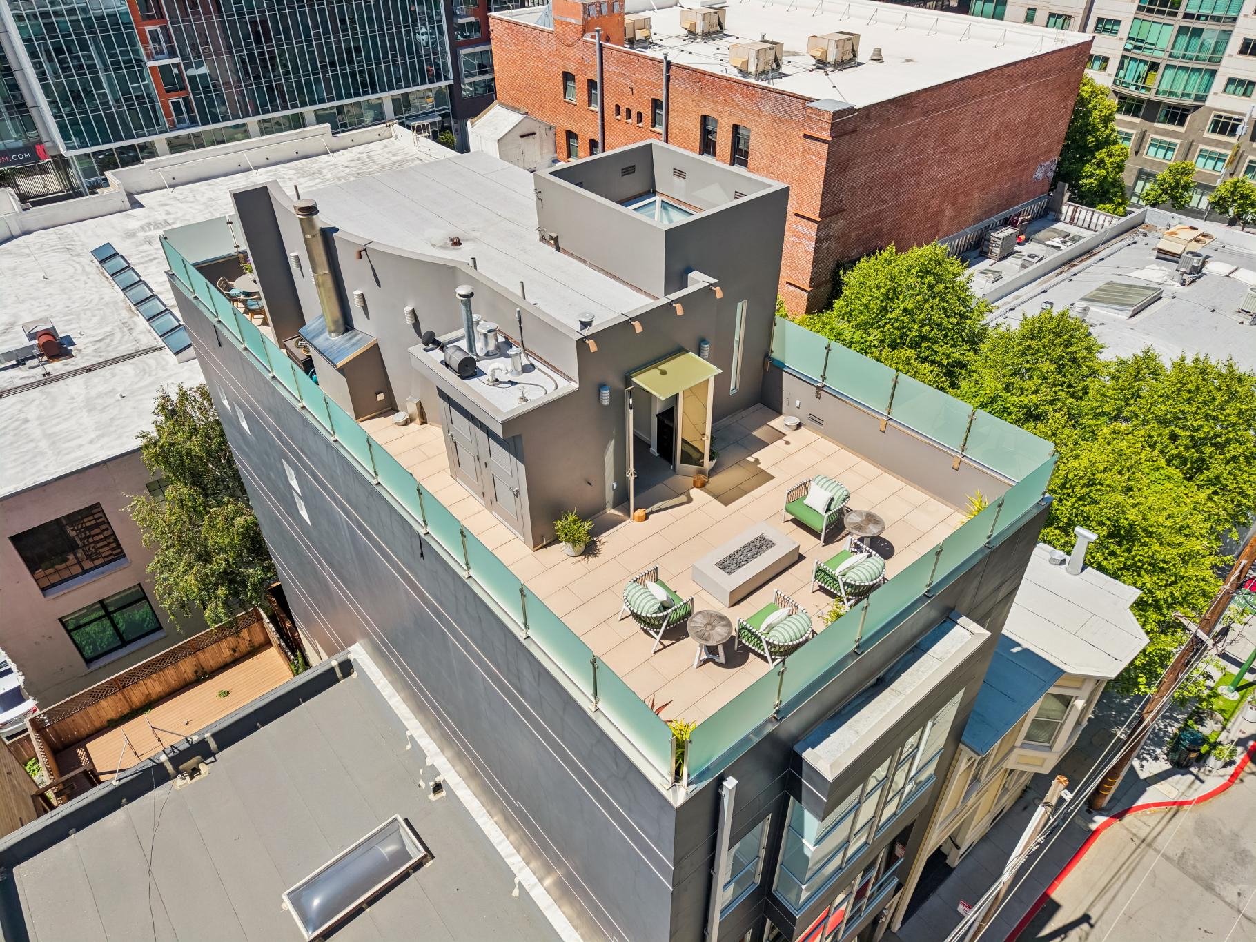 A rooftop deck on a tall building featuring outdoor furniture including chairs, tables, and a small fire pit, with a view of surrounding city buildings and trees.