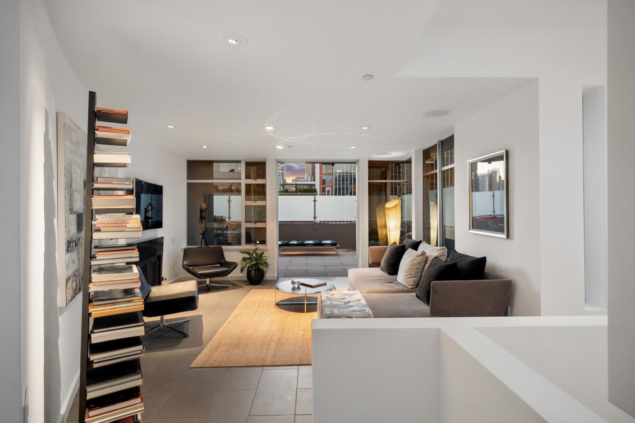 Modern living room with gray sofa, black chairs, coffee table, tall bookshelf, and large windows showing cityscape