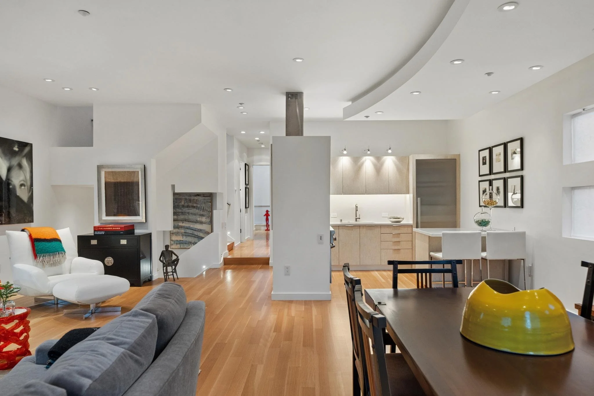 Open-concept living and dining room with white walls, hardwood floors, modern furniture, artwork, and a kitchen in the background.