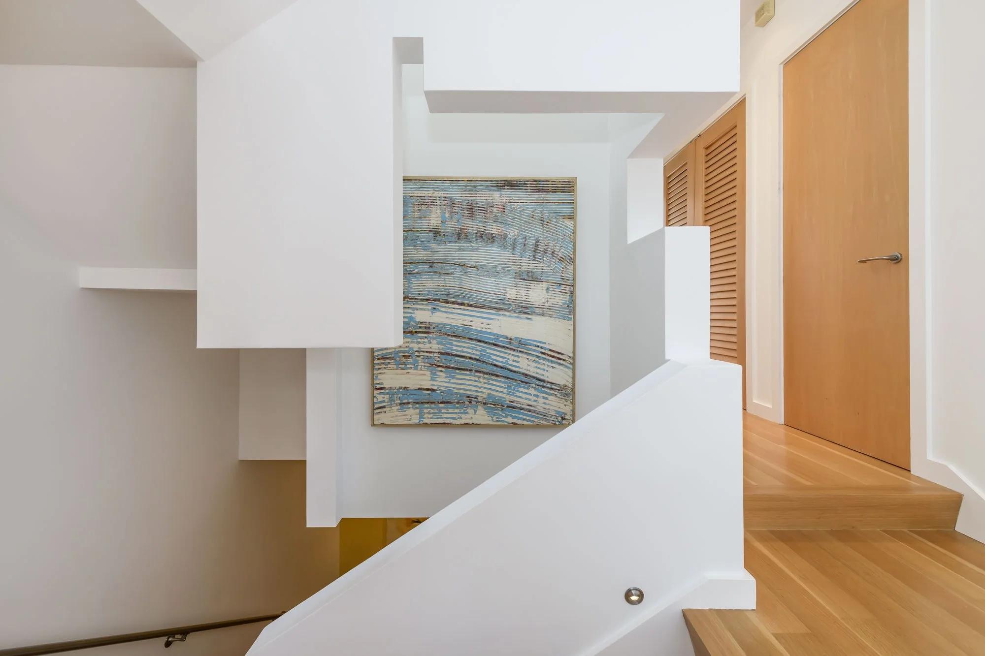 Interior of a modern house featuring a staircase with white walls, a colorful abstract painting on the wall, wooden doors, and hardwood flooring.