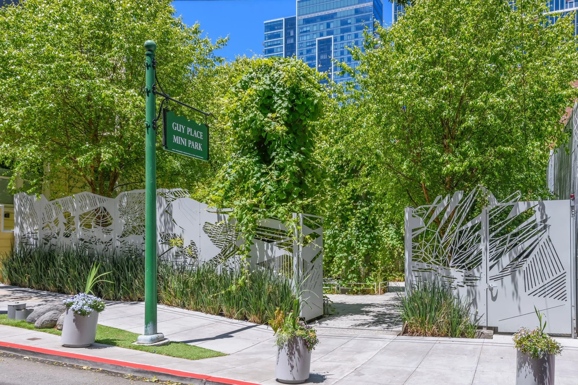 Urban park with trees, a decorative white fence, flowerpots, and a green street sign reading 'Guy Place Mini Park' with tall modern buildings in the background.