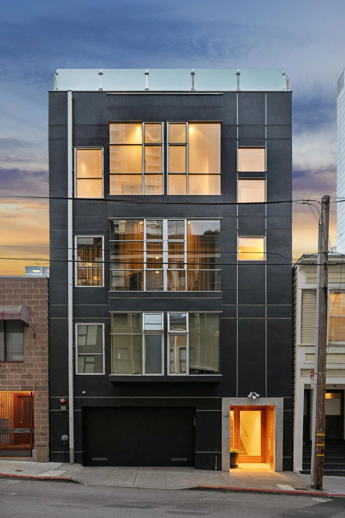A modern multi-story building with black and glass exterior, warm lighting inside, and an illuminated entrance at ground level, situated on a city street at dusk.