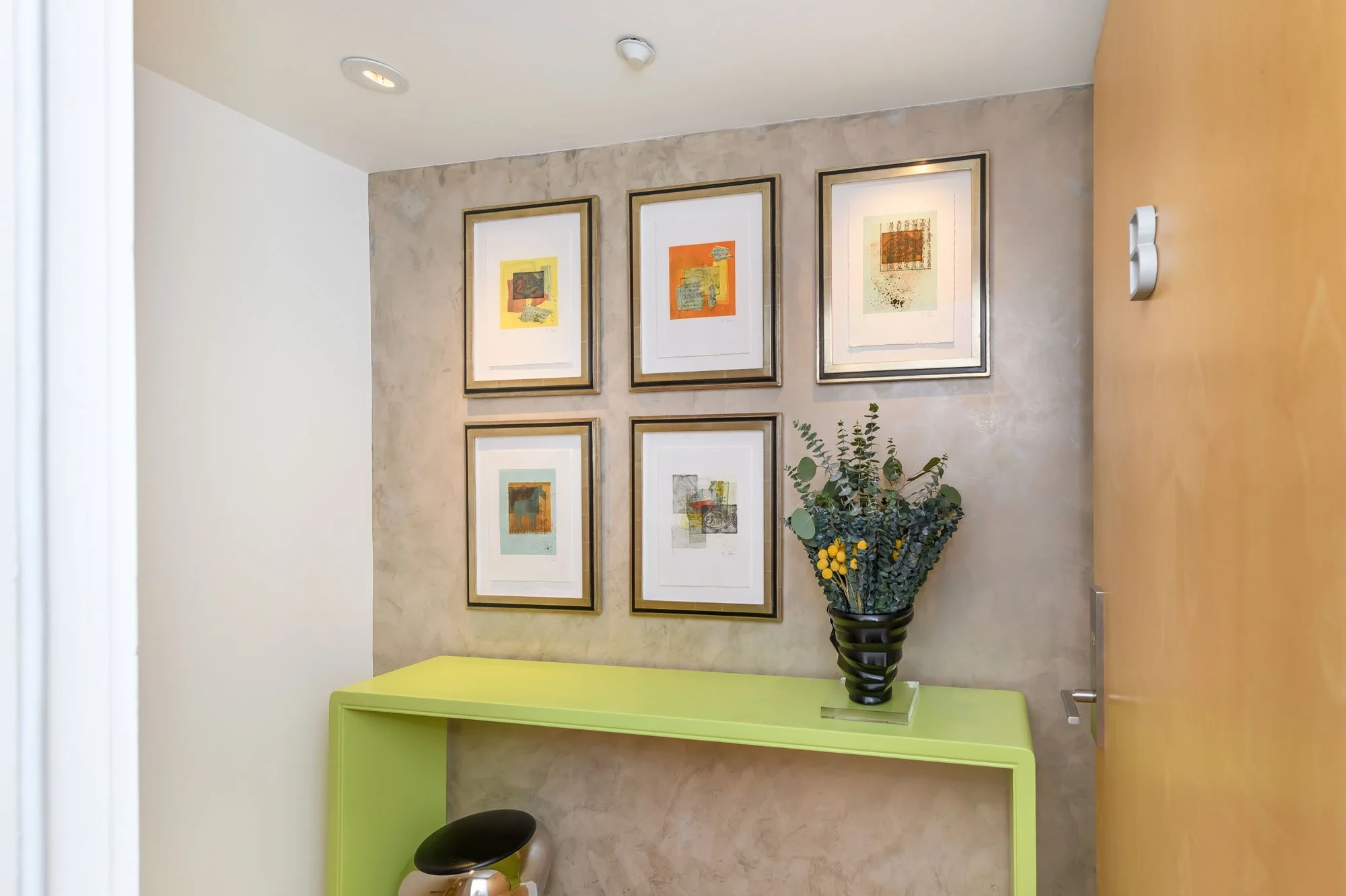 Interior view of a room with three framed abstract artwork pieces on the beige textured wall, a black vase with green and yellow foliage on a bright green table, a small black stool with a metallic base, a beige door on the right, and a white ceiling