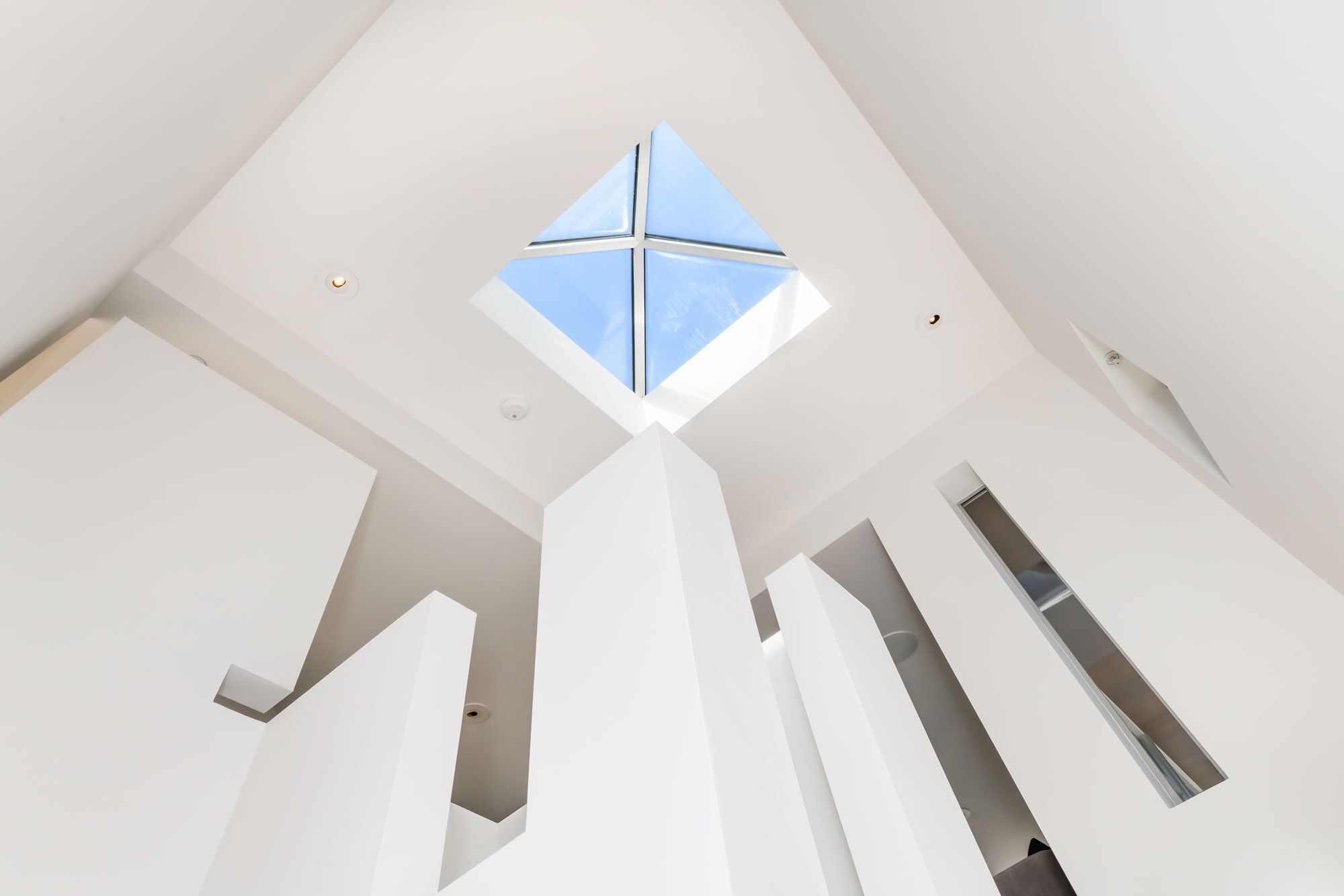 Interior view of a modern building ceiling with a square skylight letting in natural light, white walls, and some recessed ceiling lights.